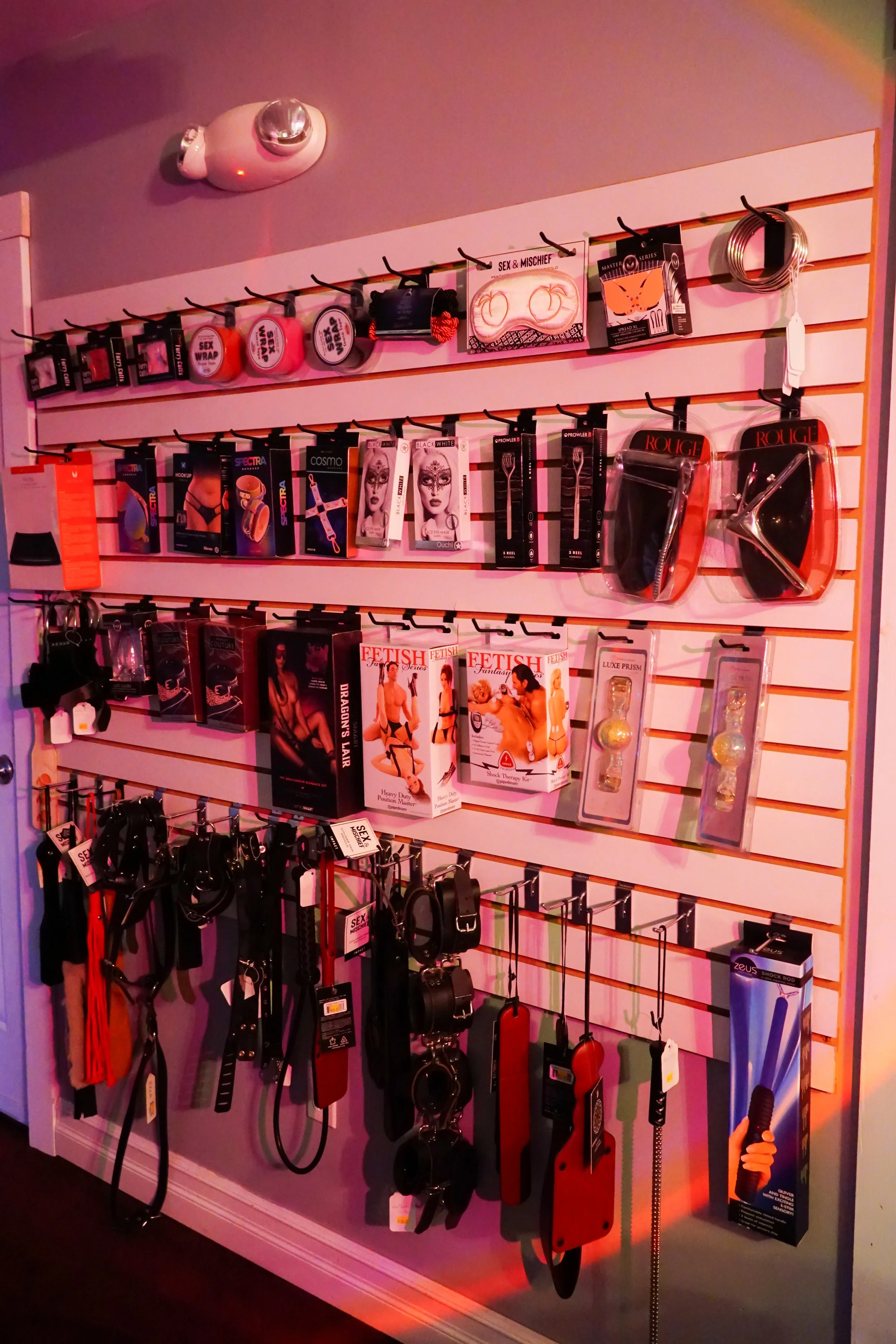 Display of adult toys and accessories, including masks, whips, cock rings, paddles, and other BDSM-related items, arranged on a wall-mounted pegboard.