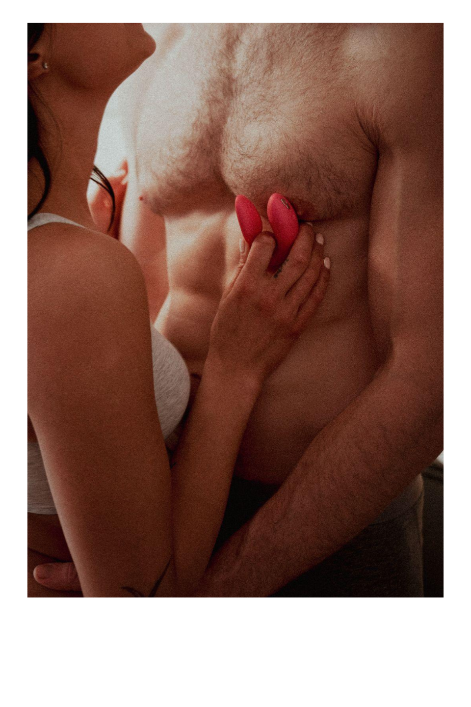 A woman with dark hair and a white top holding two pink objects close to a shirtless man's chest with chest hair, in an intimate setting.