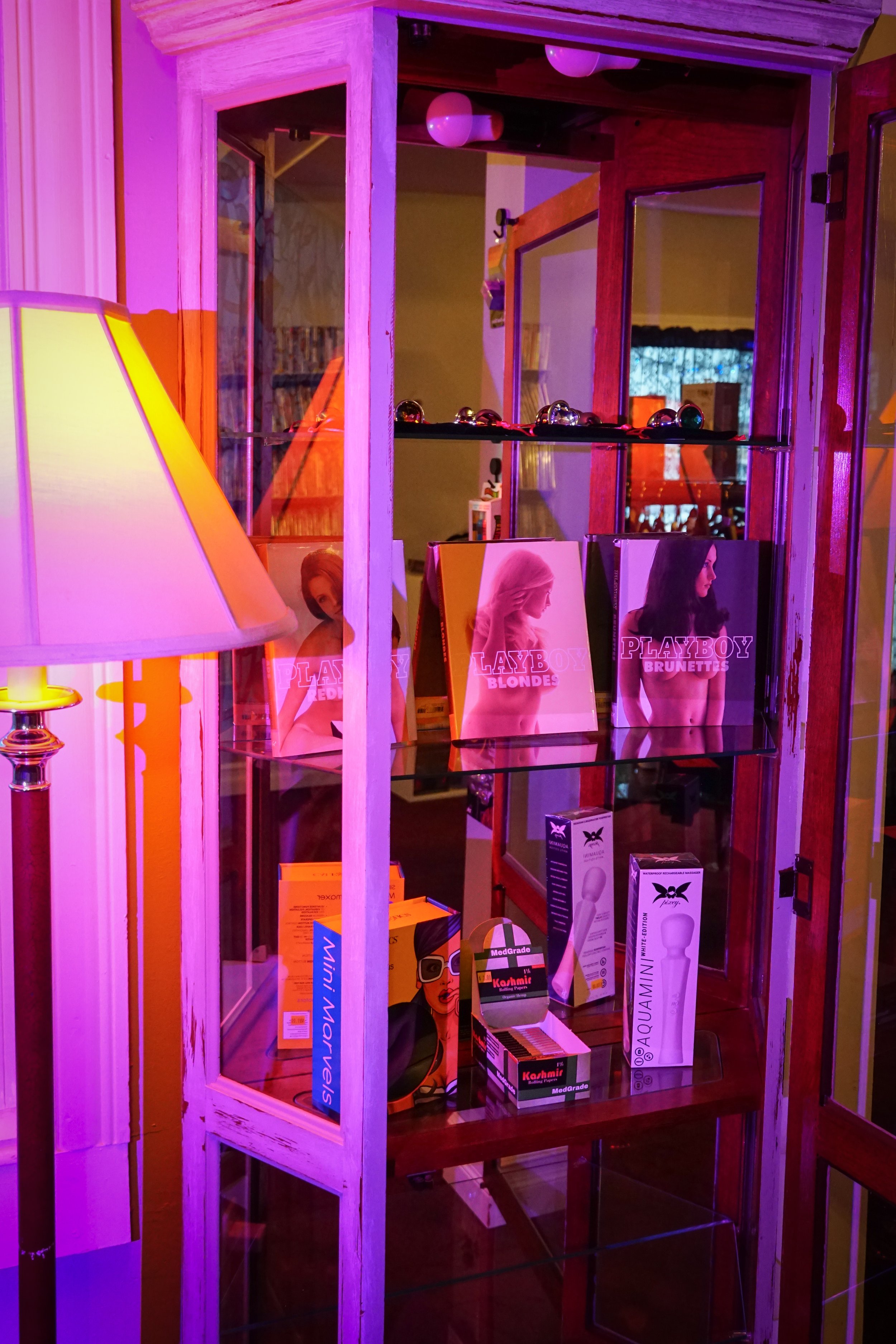 Glass display cabinet with Playboy magazines, sexual wellness products, and colorful lighting in a room with a lamp.