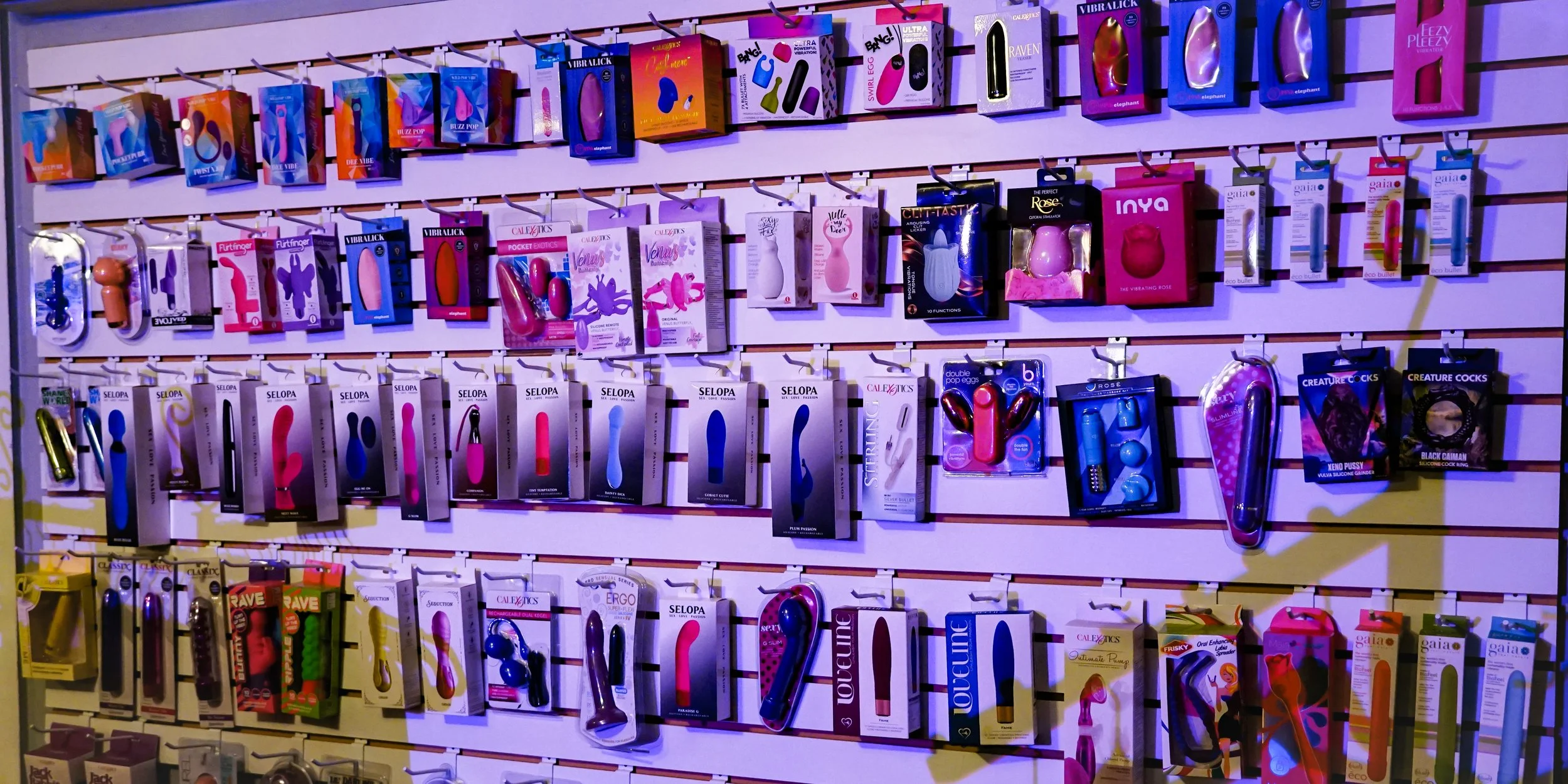 A retail display wall with various adult toys and vibrators in colorful packaging arranged on horizontal peg hooks.