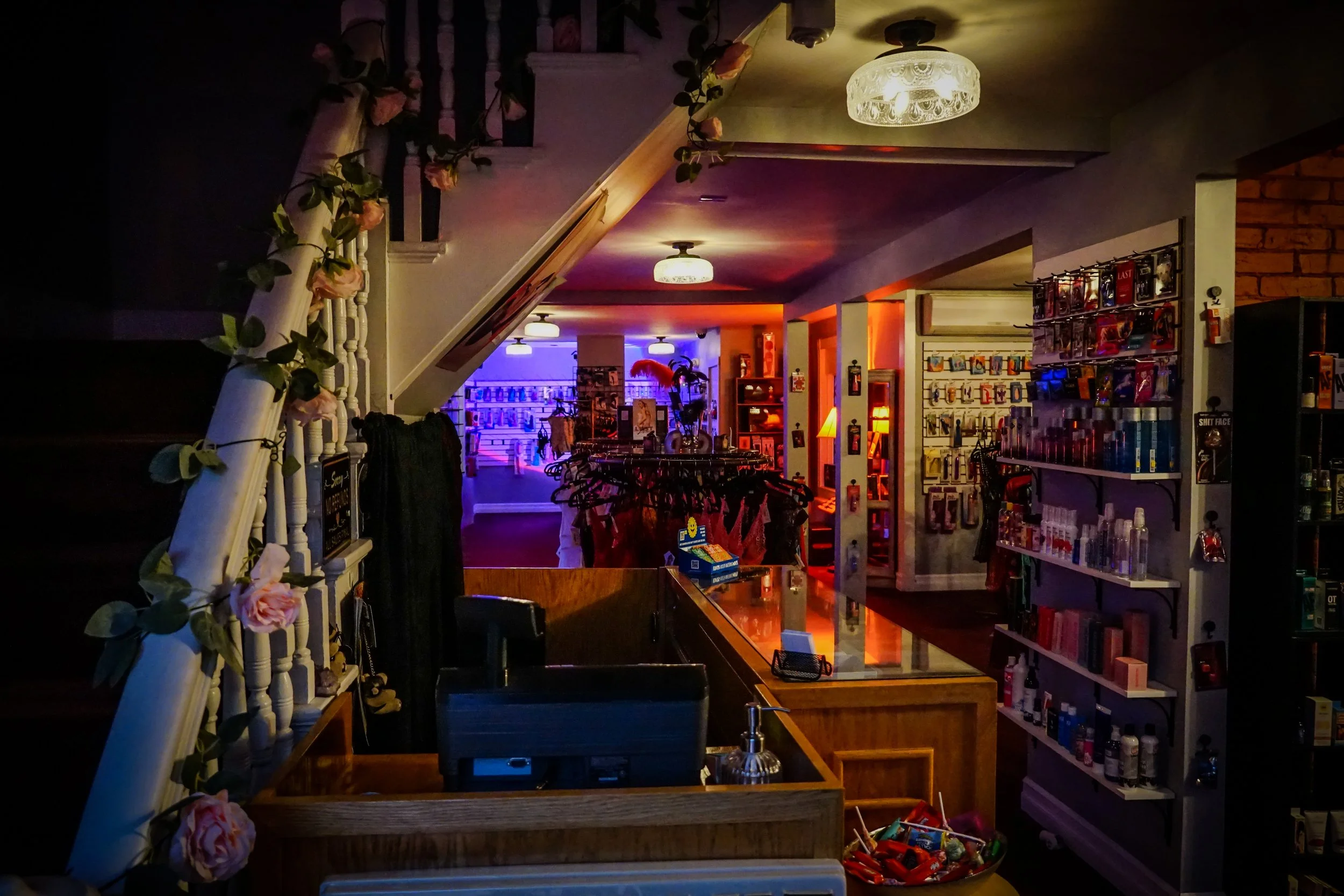 Interior of a retail store with shelves of various products, a clothing rack, a decorated staircase railing with flowers, and colorful ambient lighting in the background.