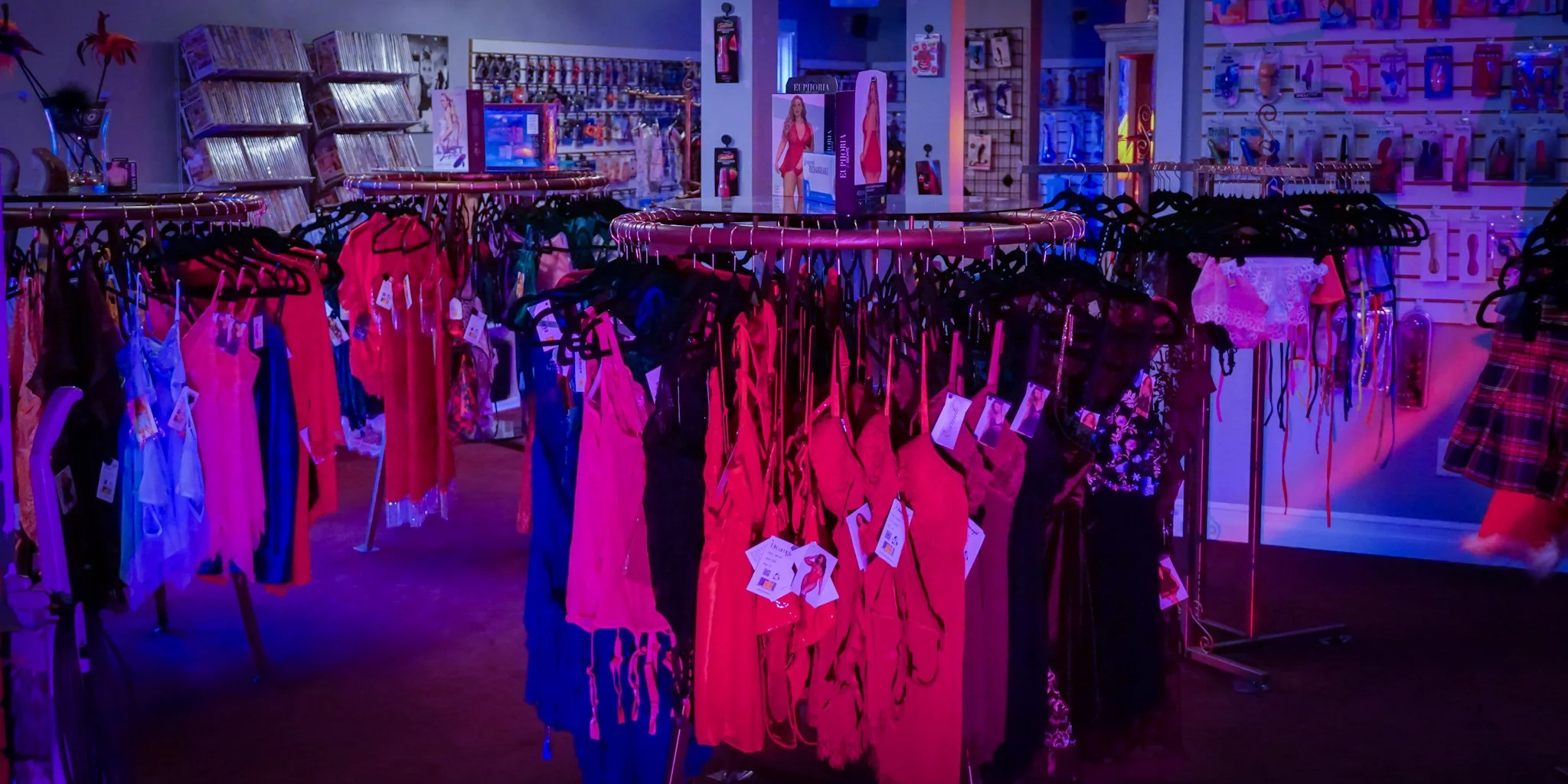 Display of colorful lingerie and sleepwear on racks inside a store with shelves of DVDs and accessories in the background, illuminated by purple and blue lighting.