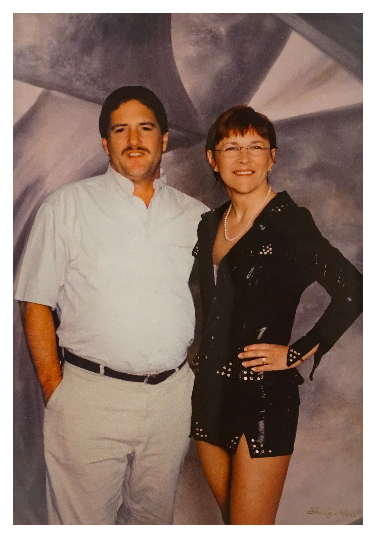 A man and a woman standing together in front of a gray abstract background. The man is wearing a white shirt and light-colored pants with a belt, and the woman is dressed in a black leather jacket and matching skirt with stud details, smiling with her hand on her hip. The woman is also wearing glasses and a pearl necklace.