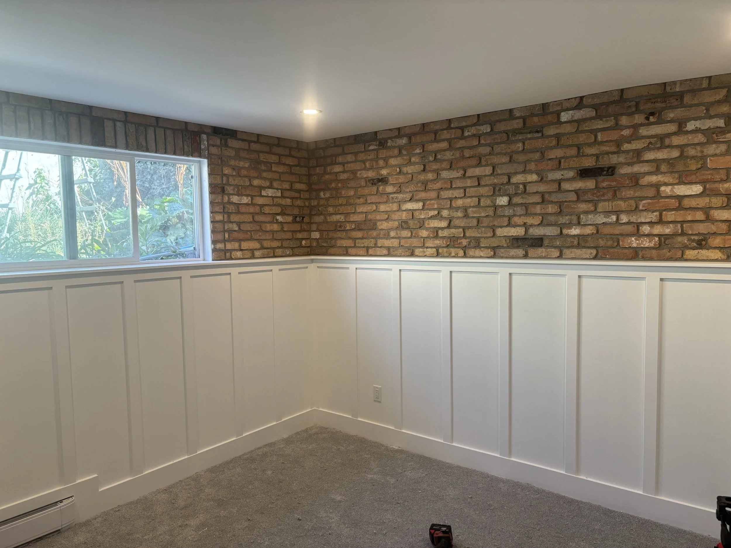 An interior room with a part brick wall, a window showing greenery outside, white paneling on the lower half of the walls, a carpeted floor, and a ceiling with recessed lighting.