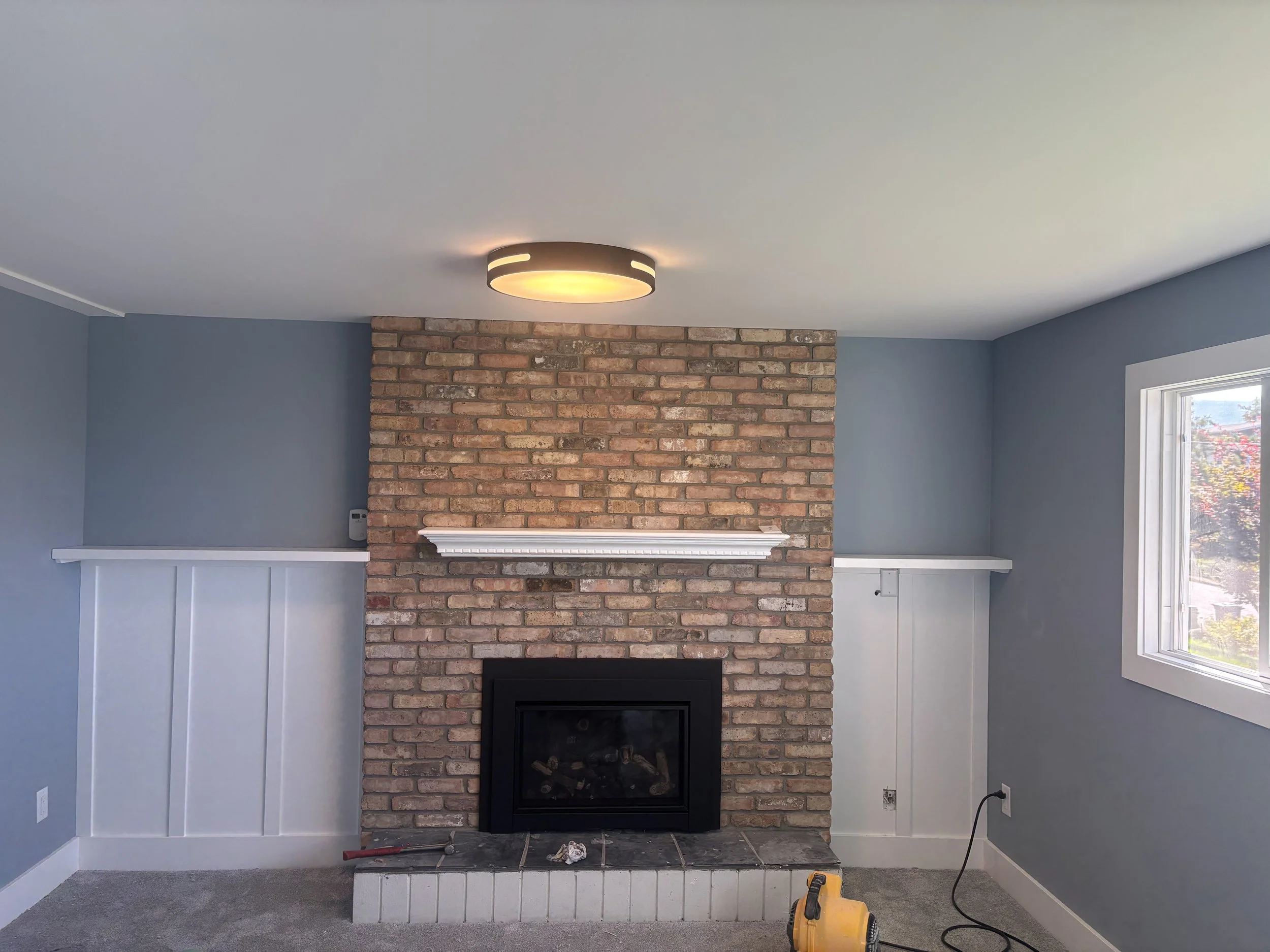 Living room with a brick fireplace, blue walls, a window on the right, and a ceiling light fixture.