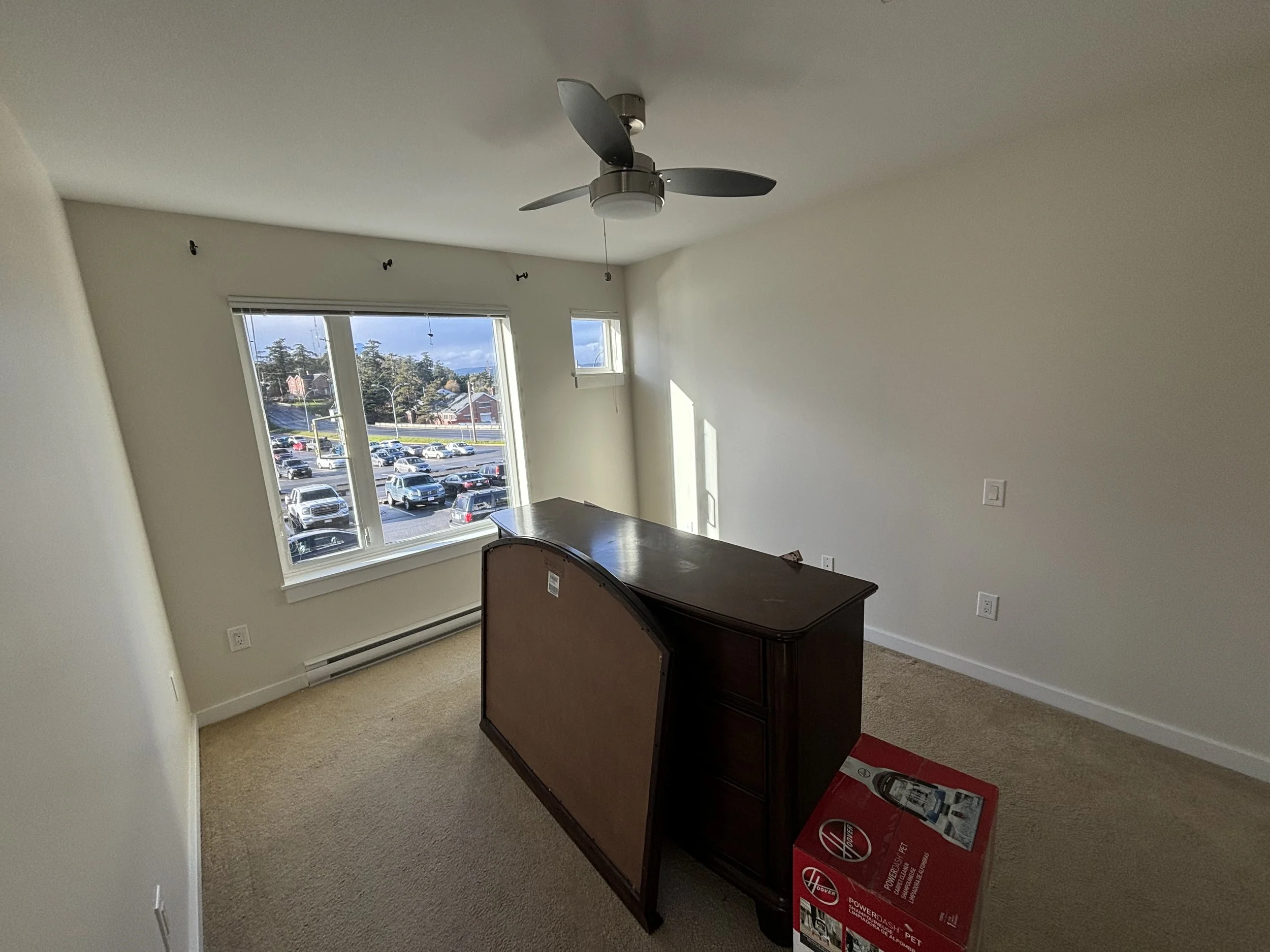 Empty room with large window overlooking a parking lot and trees, ceiling fan, beige carpet, and a wooden table with a box of Hoover PowerDash Pet carpet cleaner.