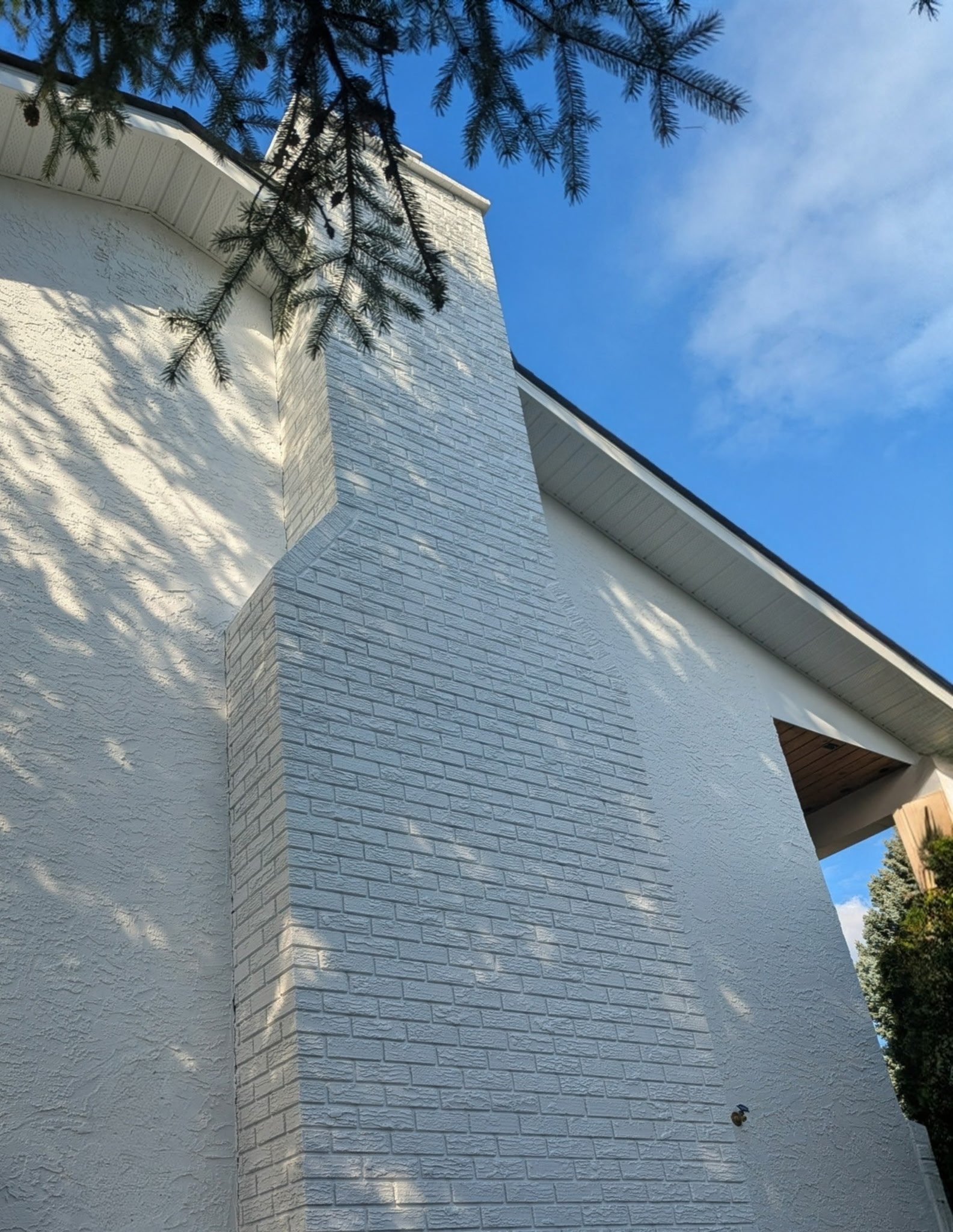 Close-up view of the corner of a white house with textured exterior walls, a chimney with a brick pattern, under a clear blue sky, with overhanging tree branches.