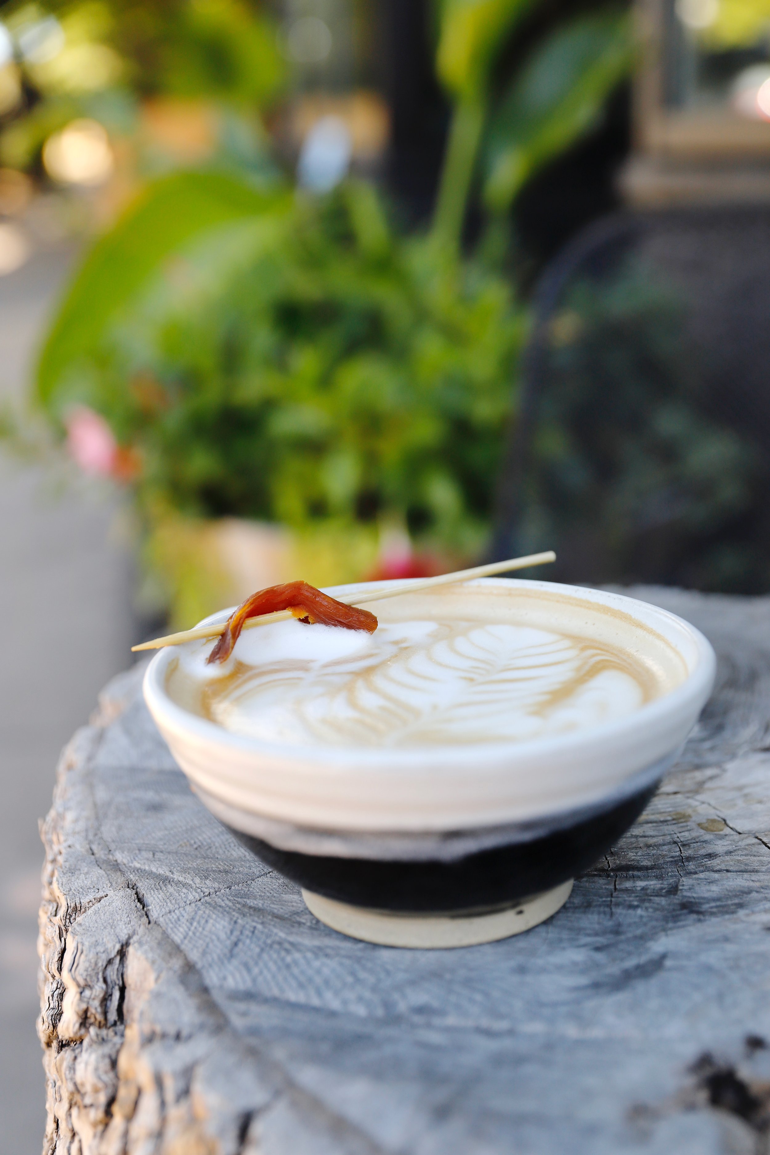 A large ceramic mug containing a specialty latte garnished with a swirl pattern, with a small sweet potato garnish on a toothpick, placed on a weathered wooden surface outdoors.