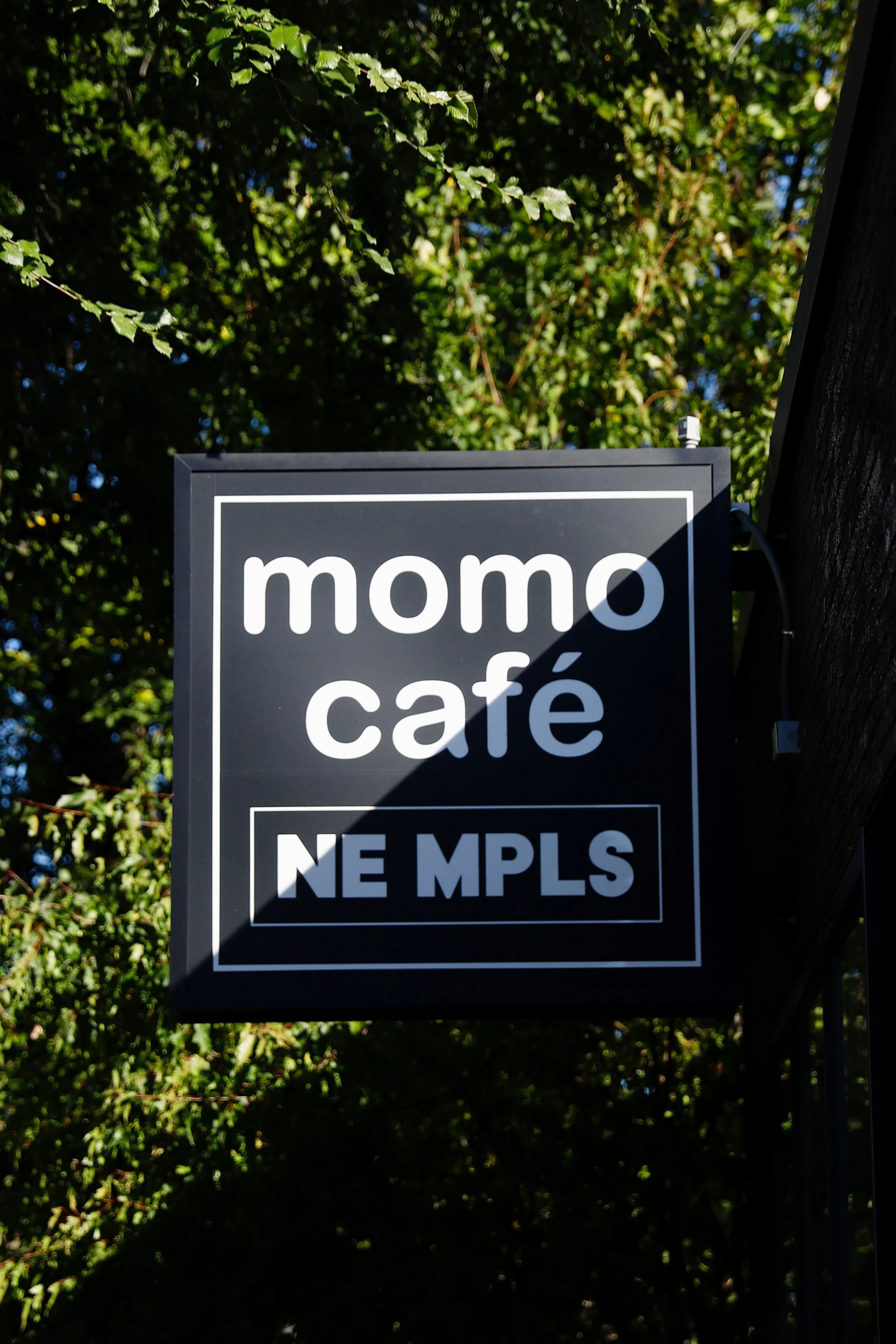 Black square sign hanging outdoors with white text reading "momo café" and "NE MPLS" below, surrounded by green trees in the background.