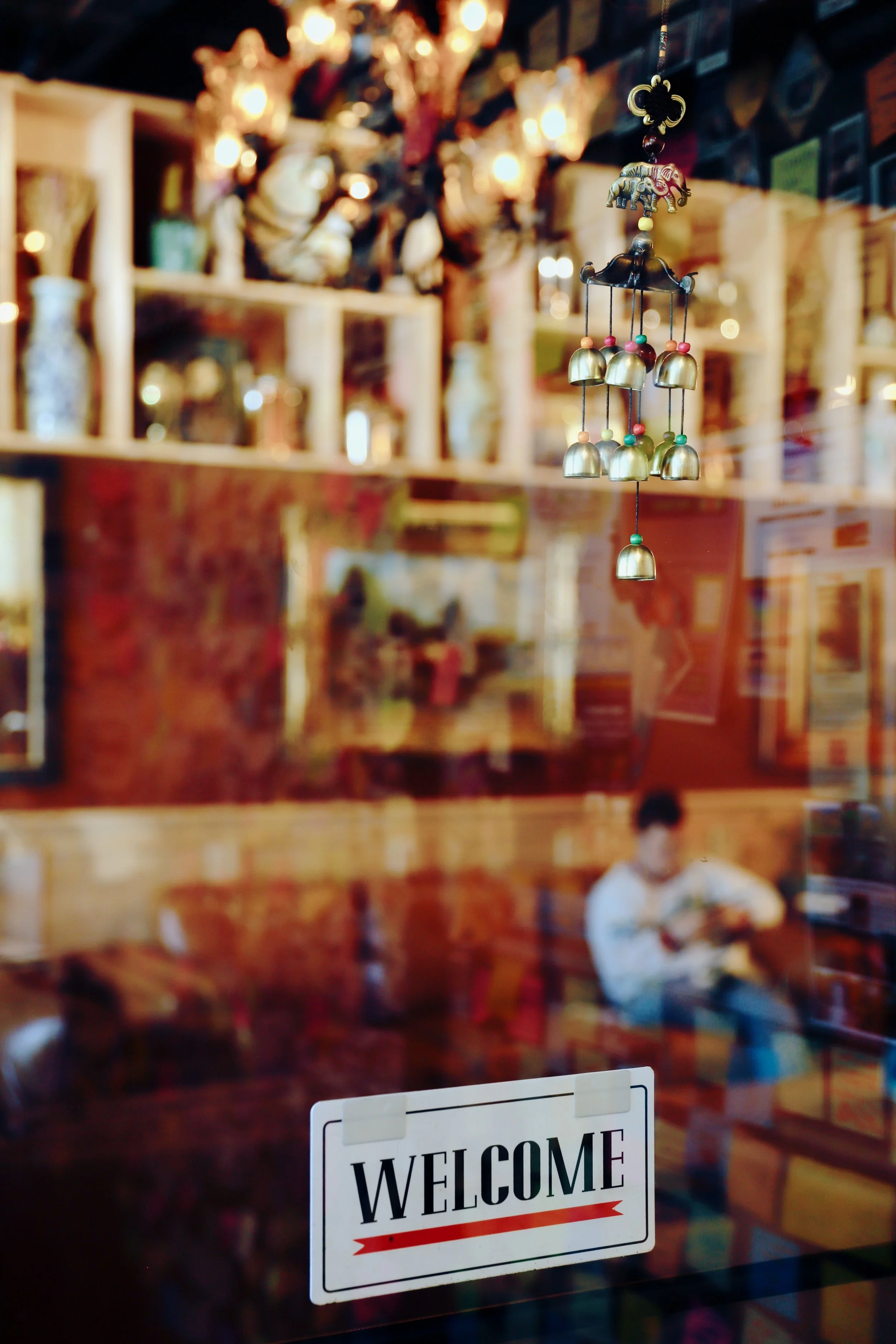 A close-up of a decorative hanging mobile with bells inside a cozy cafe, with a blurred background of shelves filled with various items and a person sitting and reading.