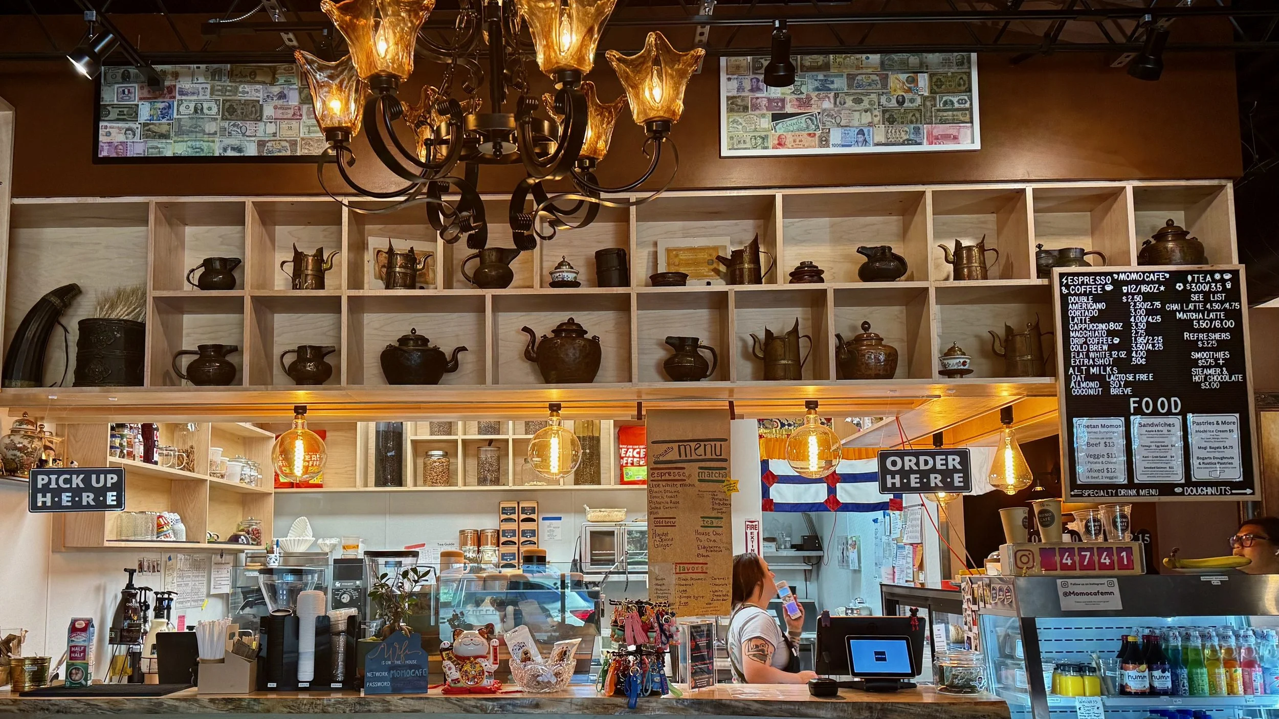 Momo Cafe interior with a wooden shelf displaying teapots and kettles, a chandelier hanging from the ceiling, a counter with a sign that says 'PICK UP HERE', a menu board, and a woman standing at the counter drinking from a cup.