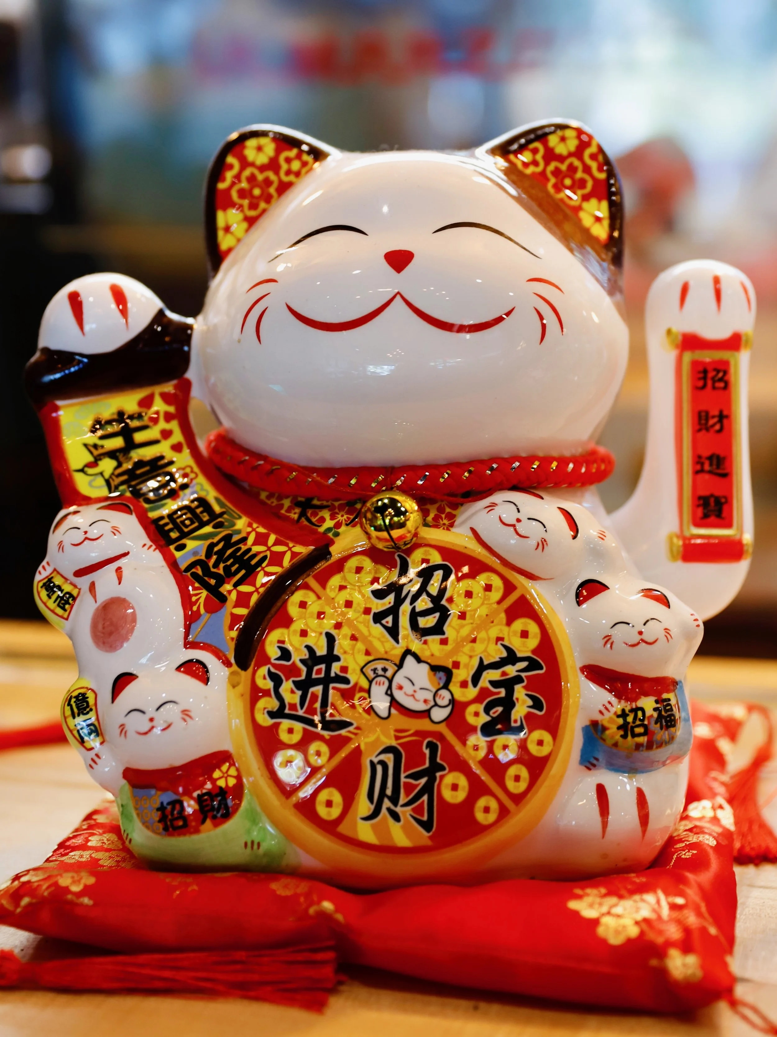A ceramic maneki-neko or lucky cat figurine with a big smiling face, holding a scroll with Chinese characters, surrounded by smaller cats, placed on red fabric.