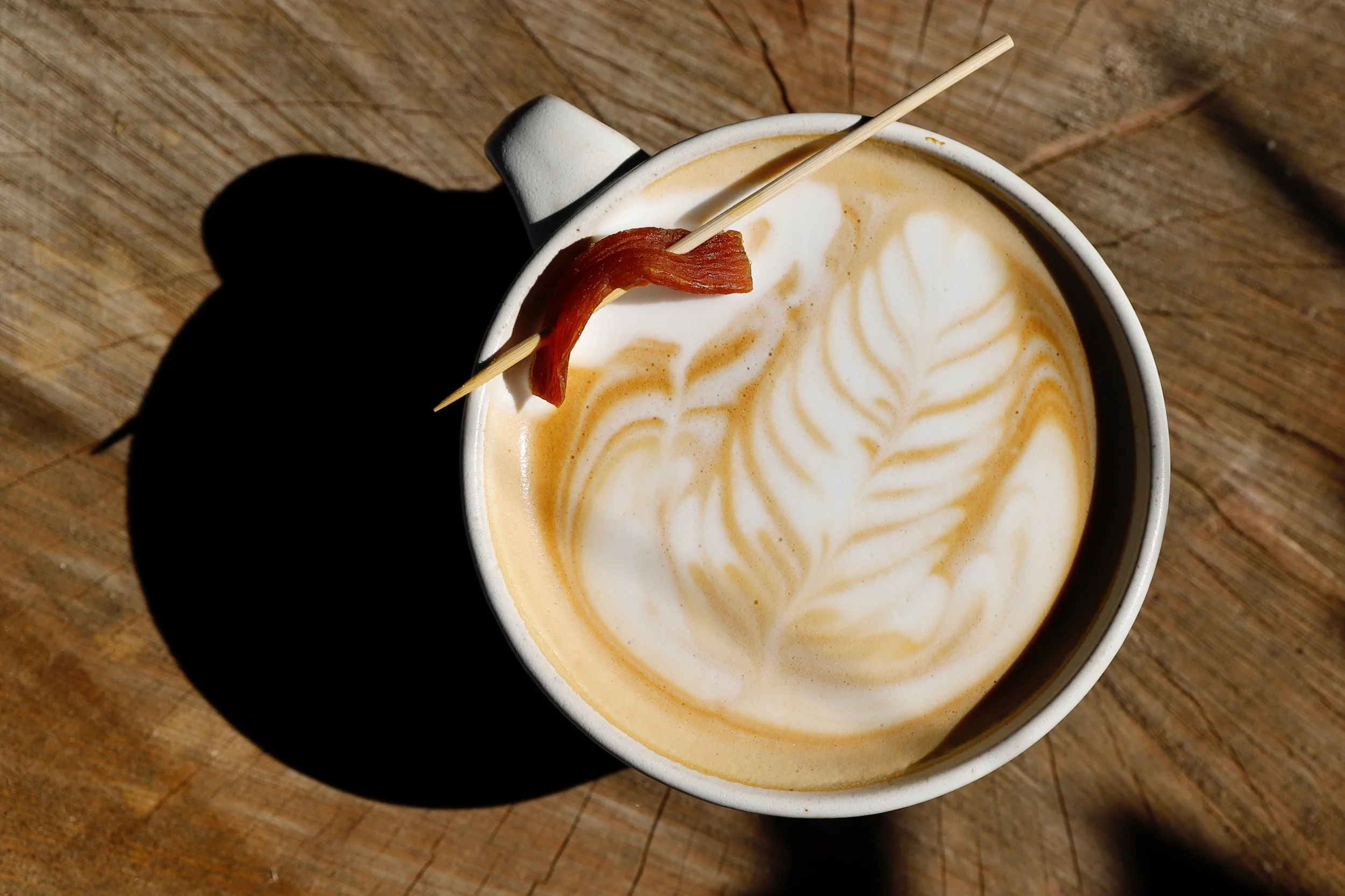 A cup of coffee with latte art and a candied sweet potato strip on a toothpick garnishing the rim, placed on a wooden surface, with a shadow cast on the left side.