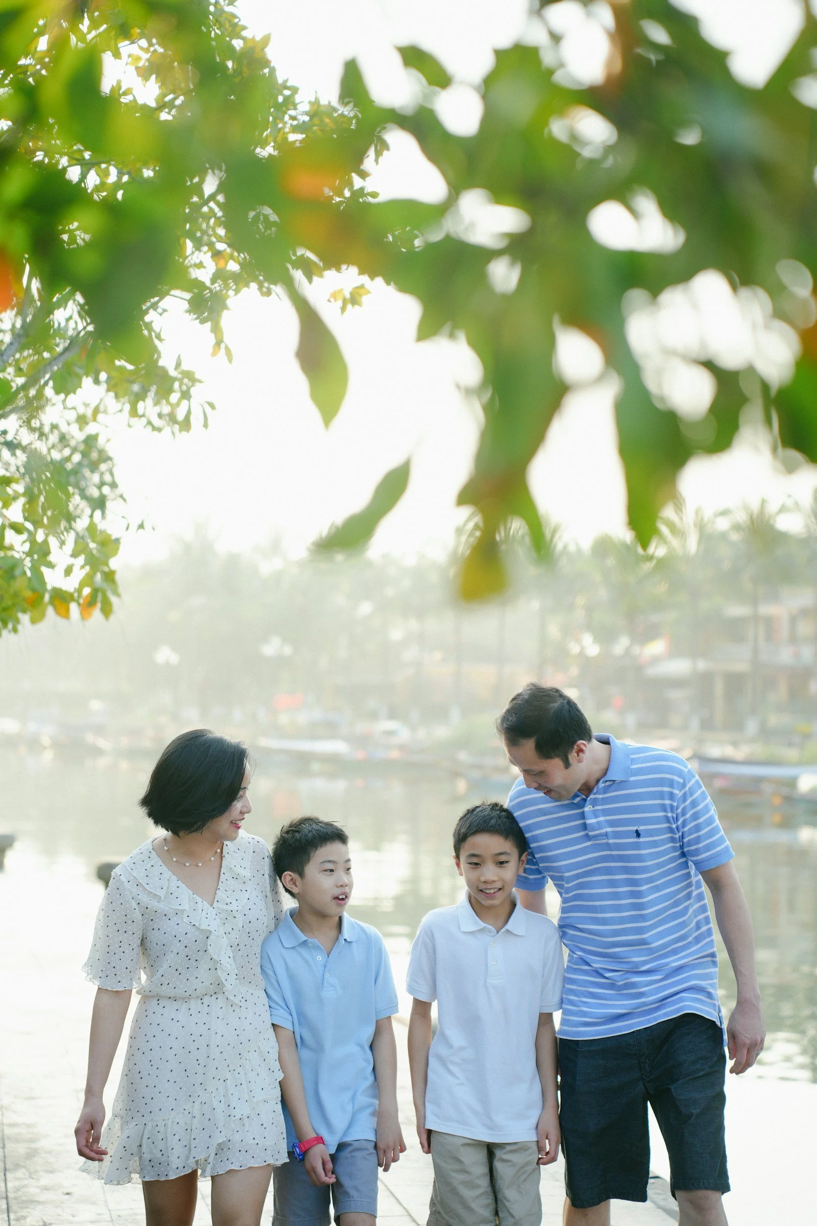 A family of four walking along a riverbank with trees overhead and boats in the water, smiling and talking together.