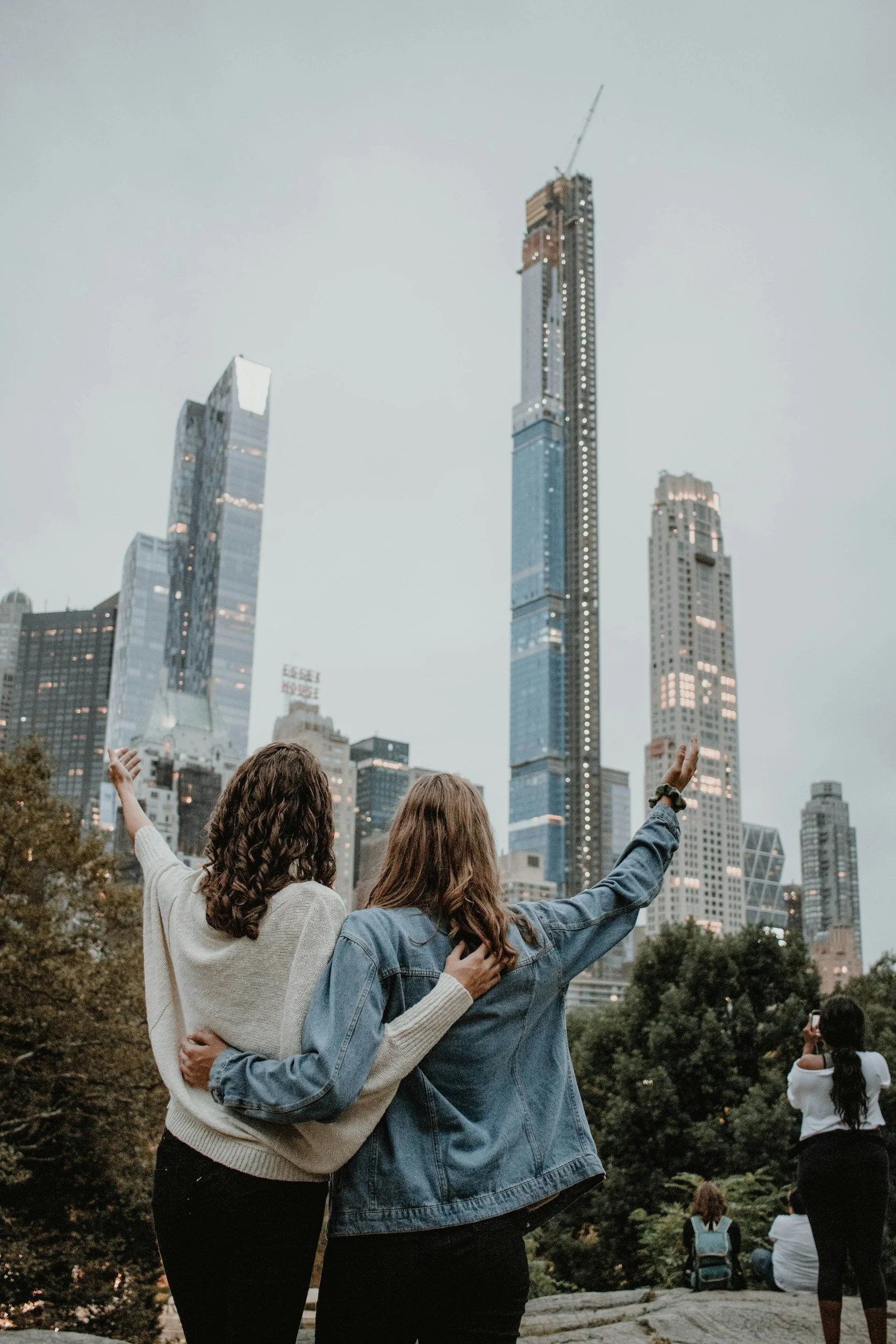 Two women standing together in a park, watching and pointing toward tall skyscrapers in a city skyline.