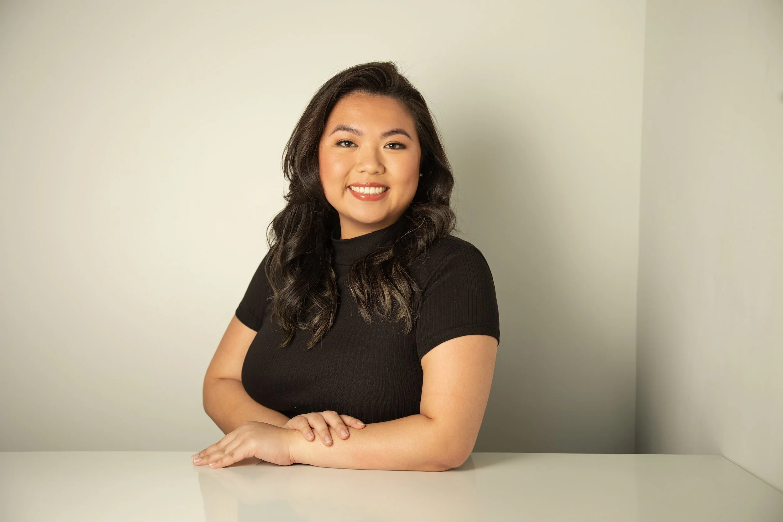 A young woman with long dark wavy hair smiling, sitting at a white table against a beige background, wearing a black short sleeve top.