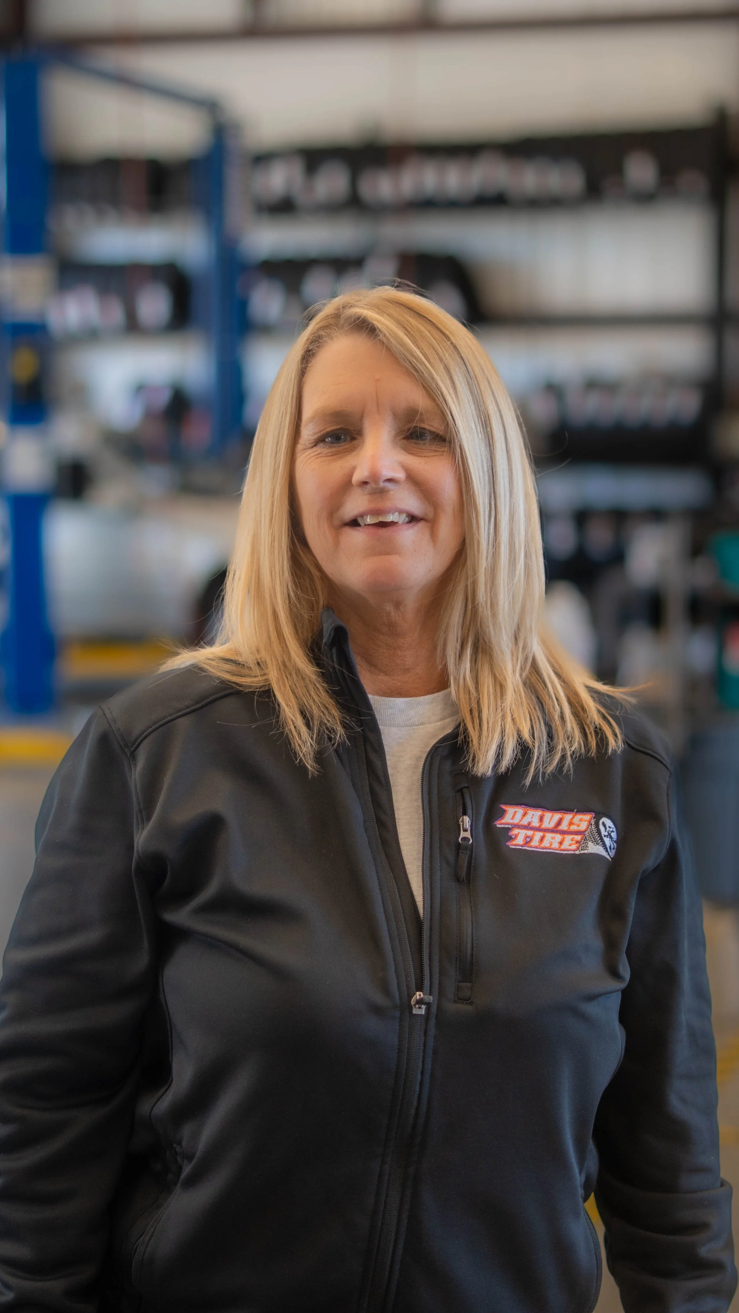 A woman with shoulder-length blonde hair smiling, wearing a black jacket with a "Davis Tire" logo, standing inside a workshop or garage with shelves full of tires and automotive supplies in the background.