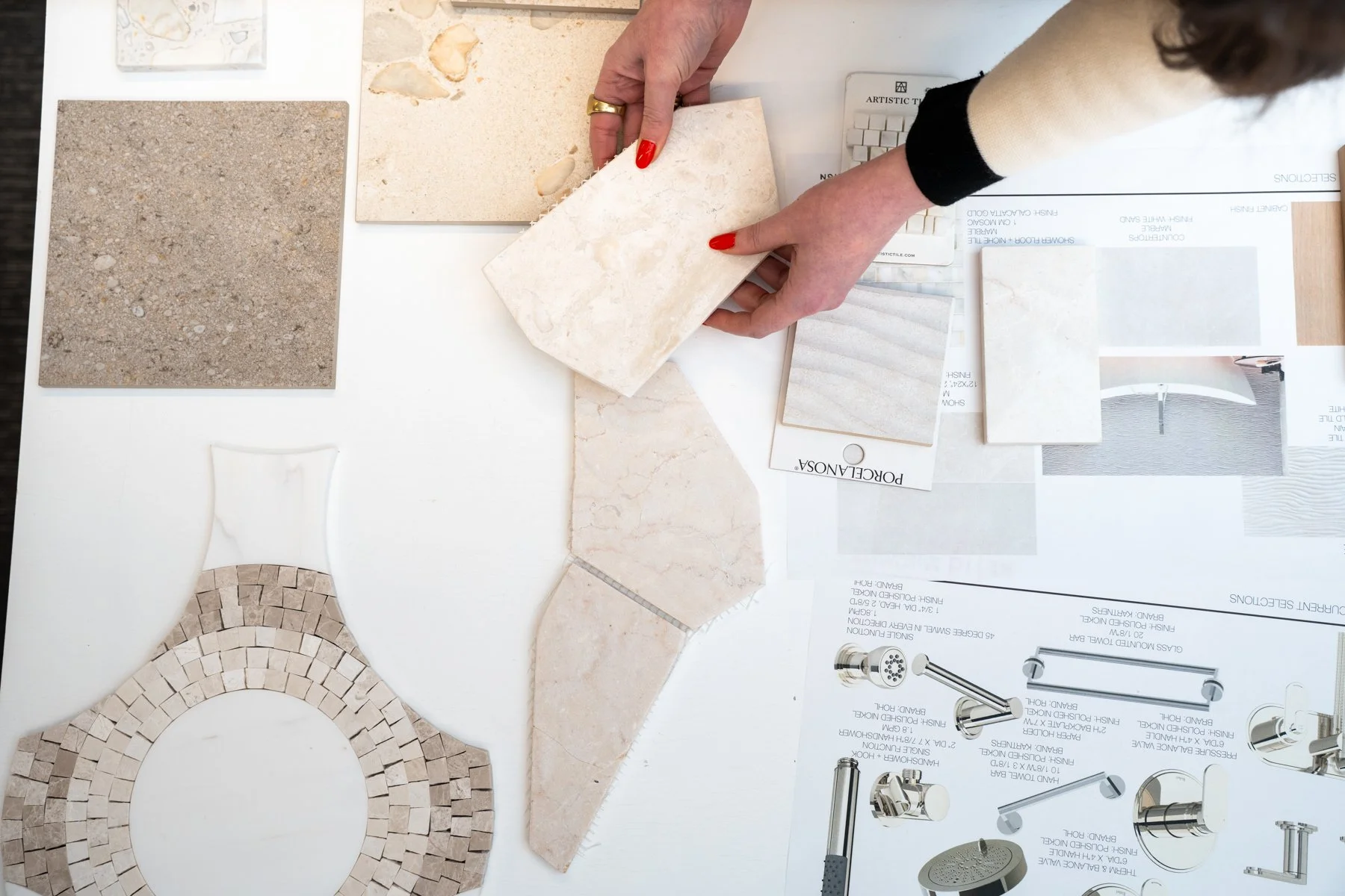A person with red nails and a gold ring is examining various sample tiles on a desk, including stone, mosaic, and porcelain options, along with a brochure and material samples for interior design or renovation planning.