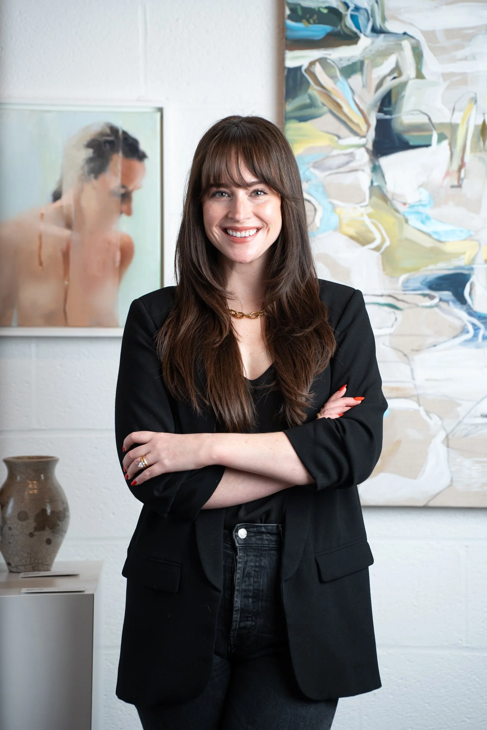Julia Machado, Interior Designer and Project Manager, smiling with crossed arms, wearing a black blazer and gold necklace, standing in front of abstract paintings and a pottery vase.