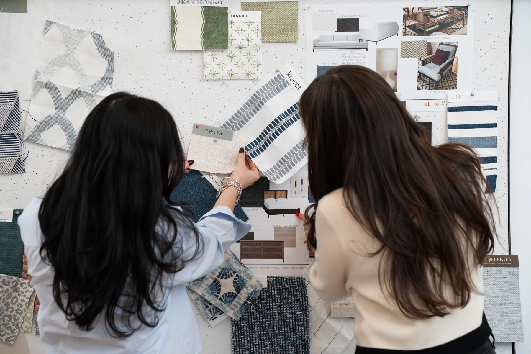 Two women are looking at fabric swatches and design samples pinned on a white board for interior decorating.