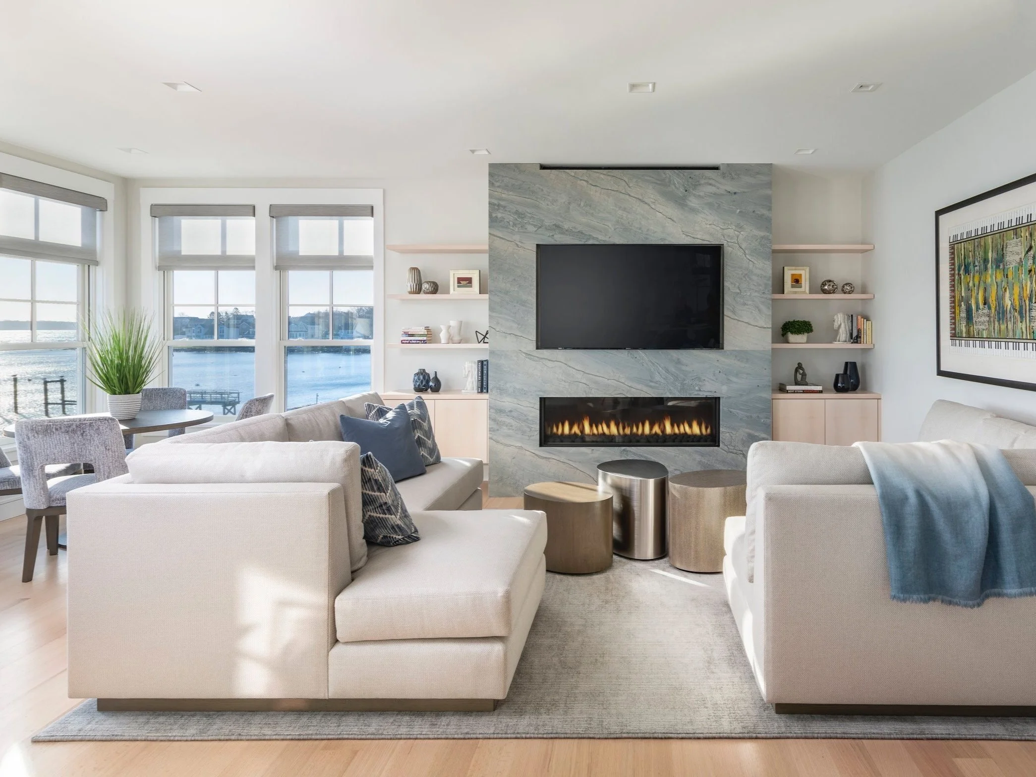 A modern living room with large windows overlooking water, white sofas, a fireplace with a marble surround, and shelves with decorative items and books.