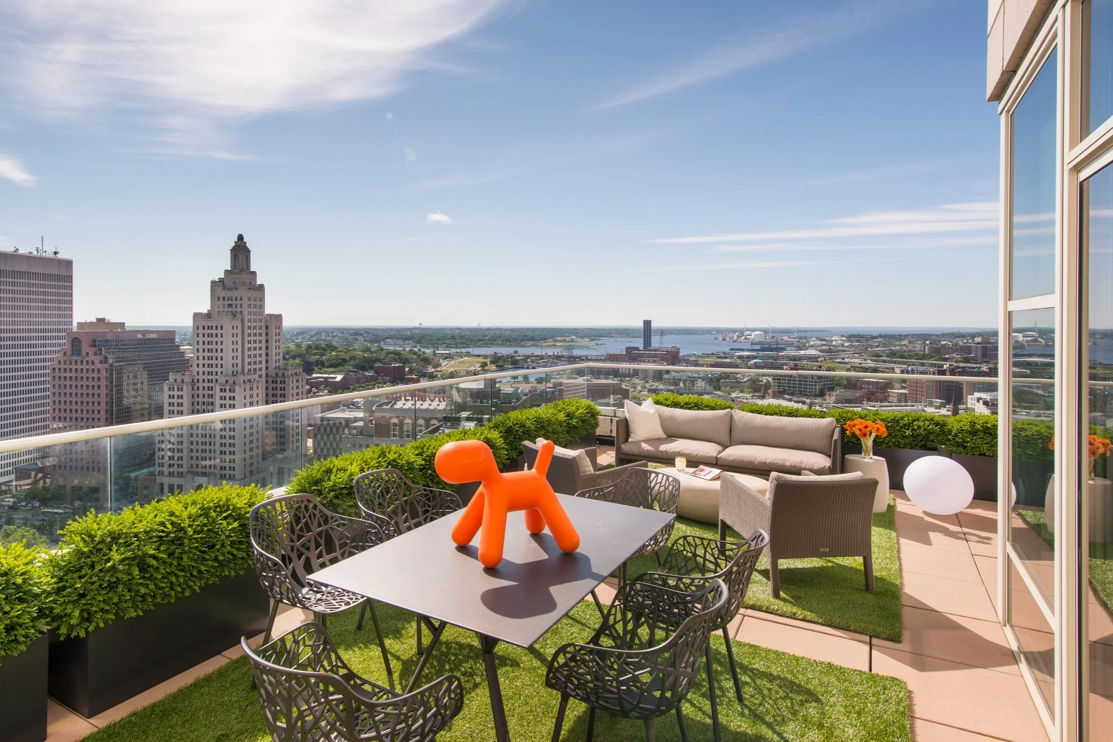 City skyline view from a balcony with outdoor furniture, including a metal table with black chairs, a white sofa, and potted flowers, overlooking tall buildings, a river, and blue sky with clouds.