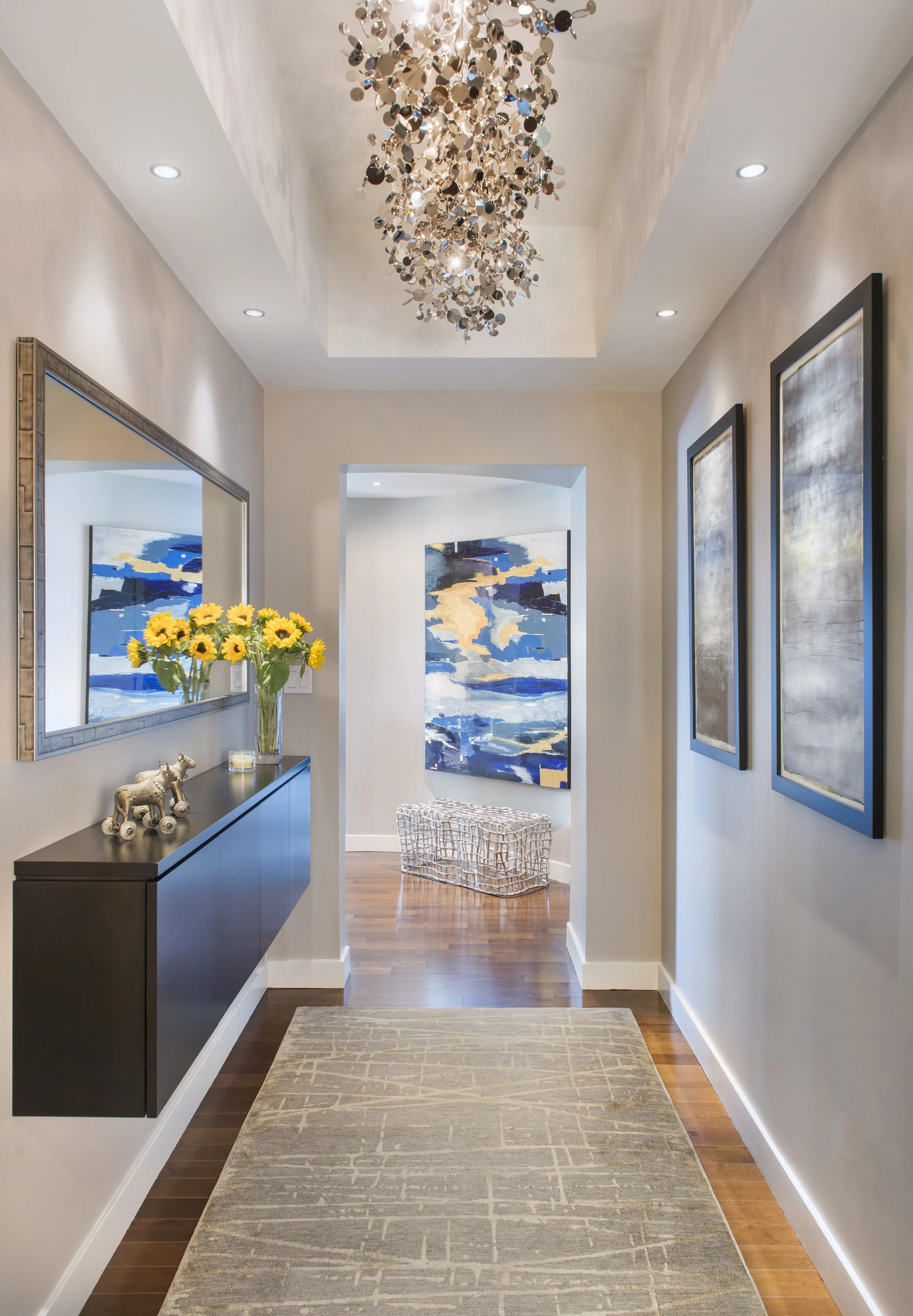 An interior hallway with beige walls, wooden flooring, and a beige rug. There is a hallway table on the left with a mirror above it, a bouquet of yellow sunflowers, and decorative figurines. Recessed ceiling lights illuminate the space. At the end, the hallway opens into a room with abstract blue and beige artwork and a white wire side table.