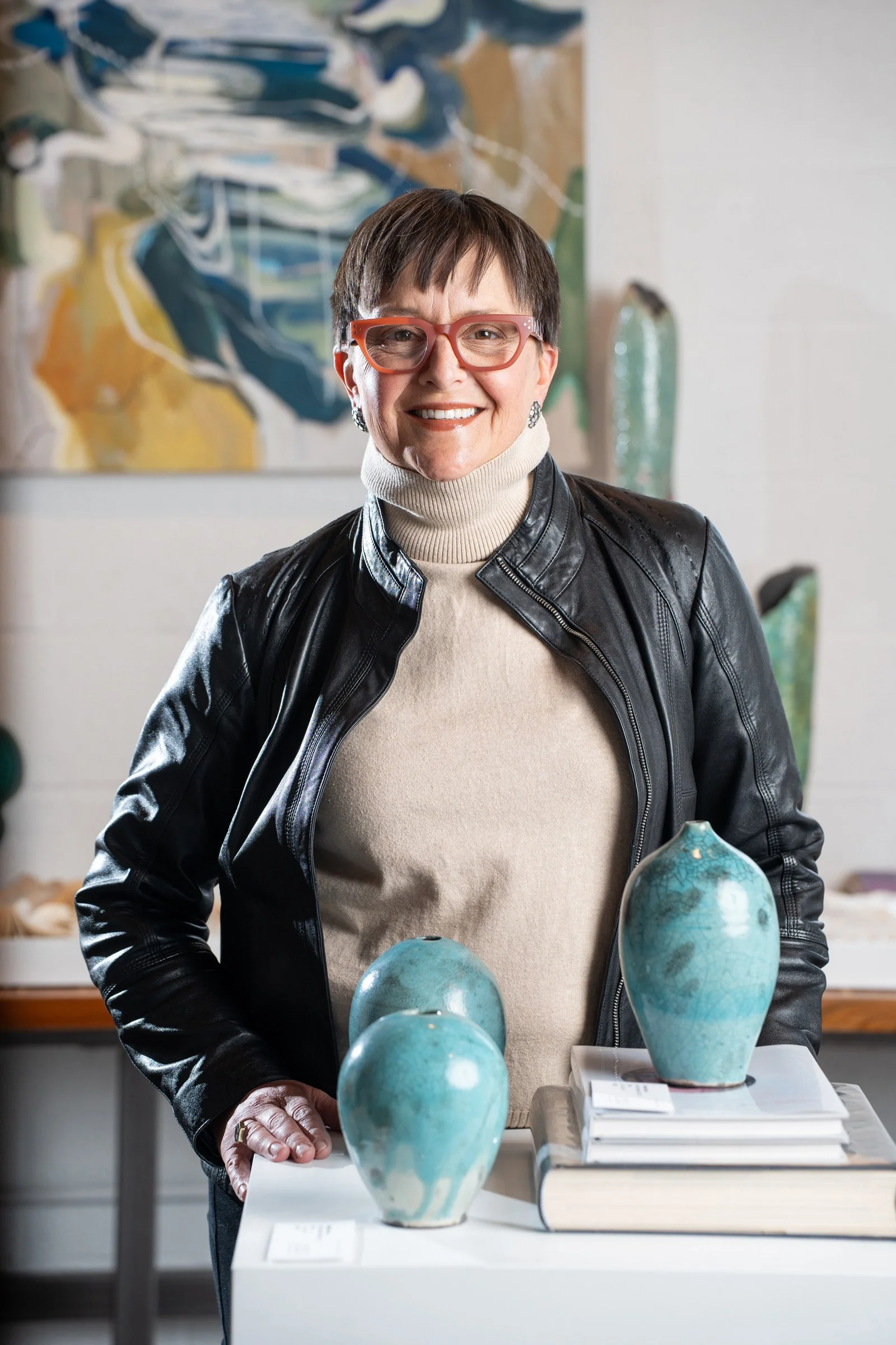  Kelly Taylor, NCIDQ, IIDA, LEED GA, Principal and Creative Director, with short dark hair, wearing red glasses, a beige turtleneck, and a black leather jacket, standing in front of an abstract painting.