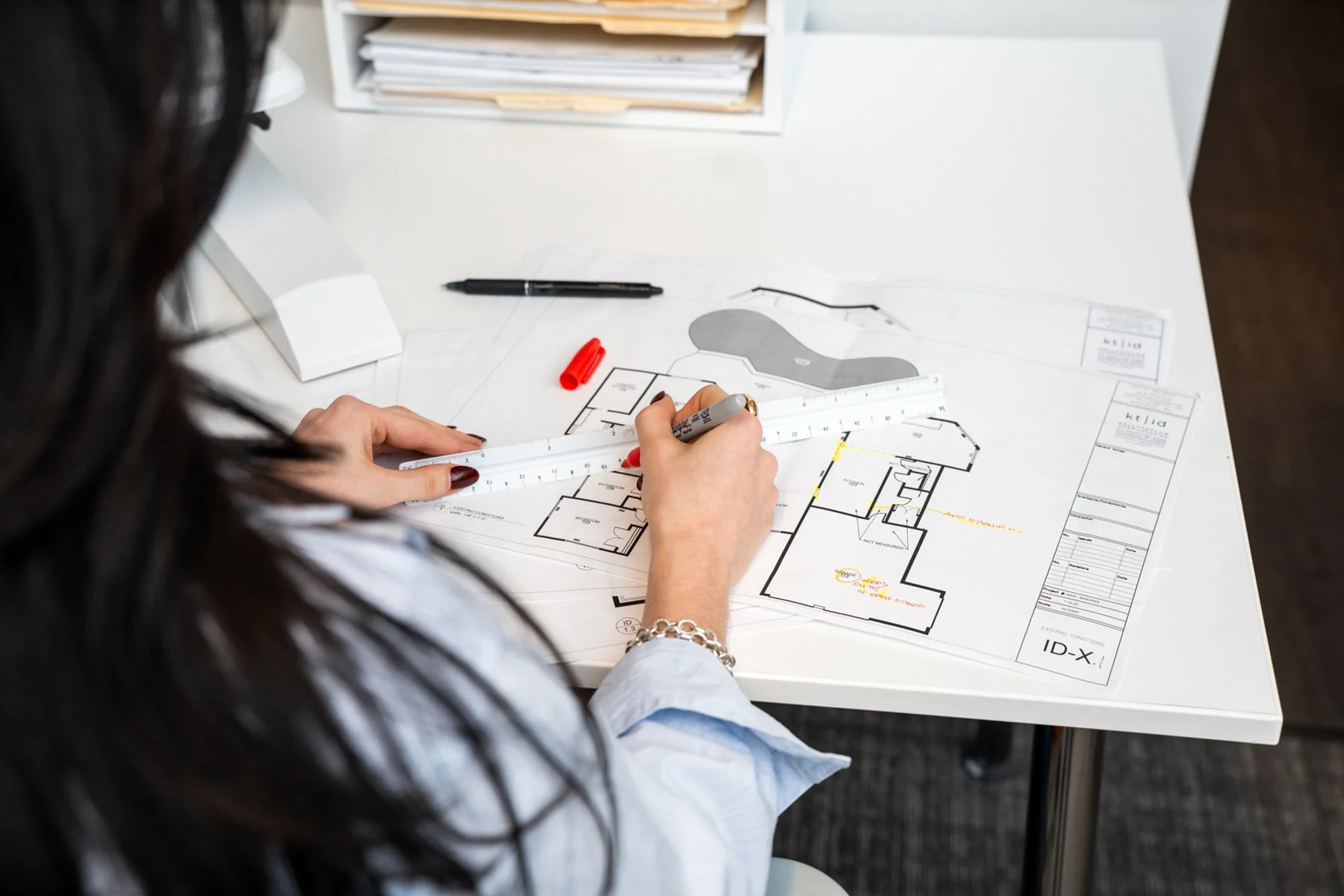 A person reviewing architectural floor plans and blueprints at a desk, using a ruler to measure the plans, with pens and markers nearby.