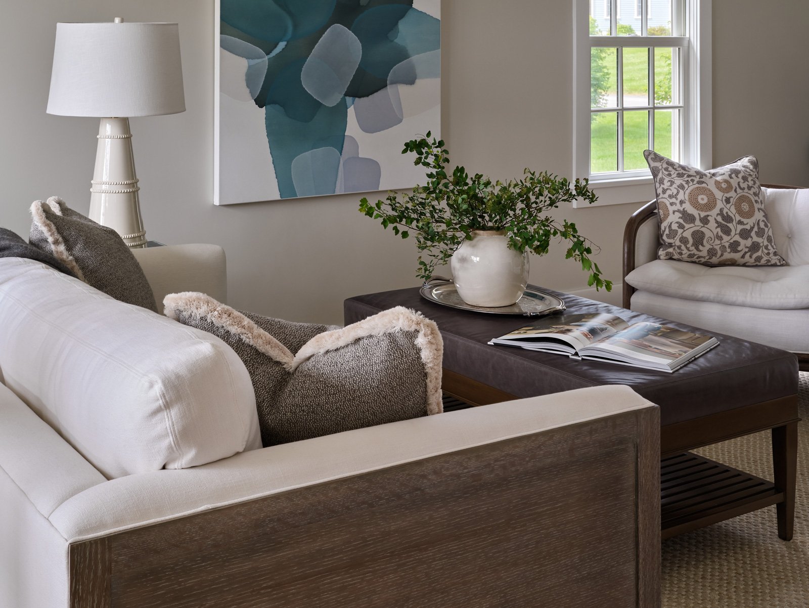 Living room with white sofas, a dark brown coffee table with a book and a vase with green foliage, a cream-colored table lamp, a modern abstract painting, and a window with a view of a green lawn.
