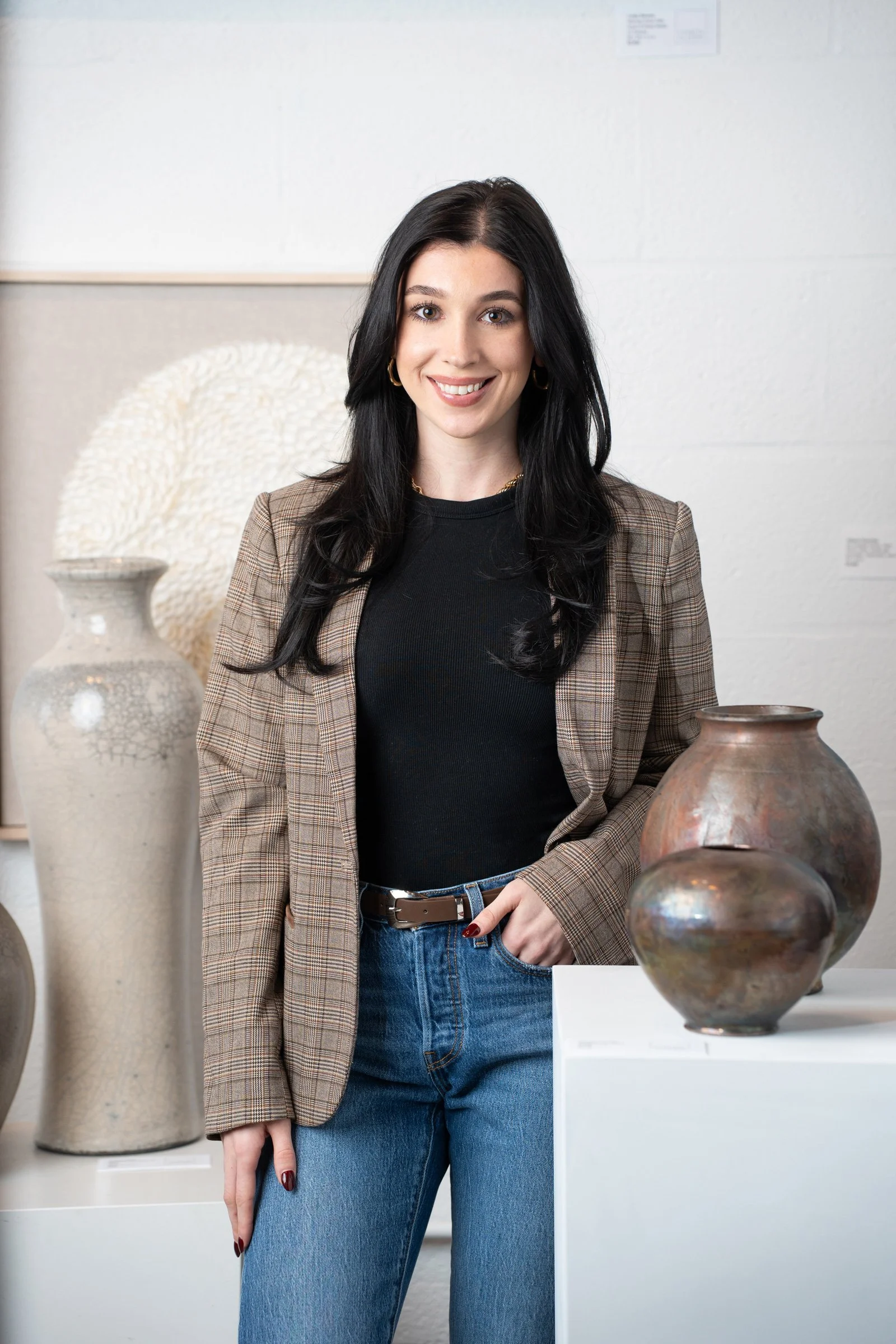 Madison Famolle, Assistant Designer, smiling at the camera, wearing a black top, plaid blazer, and blue jeans, standing next to pottery vases in an art gallery.
