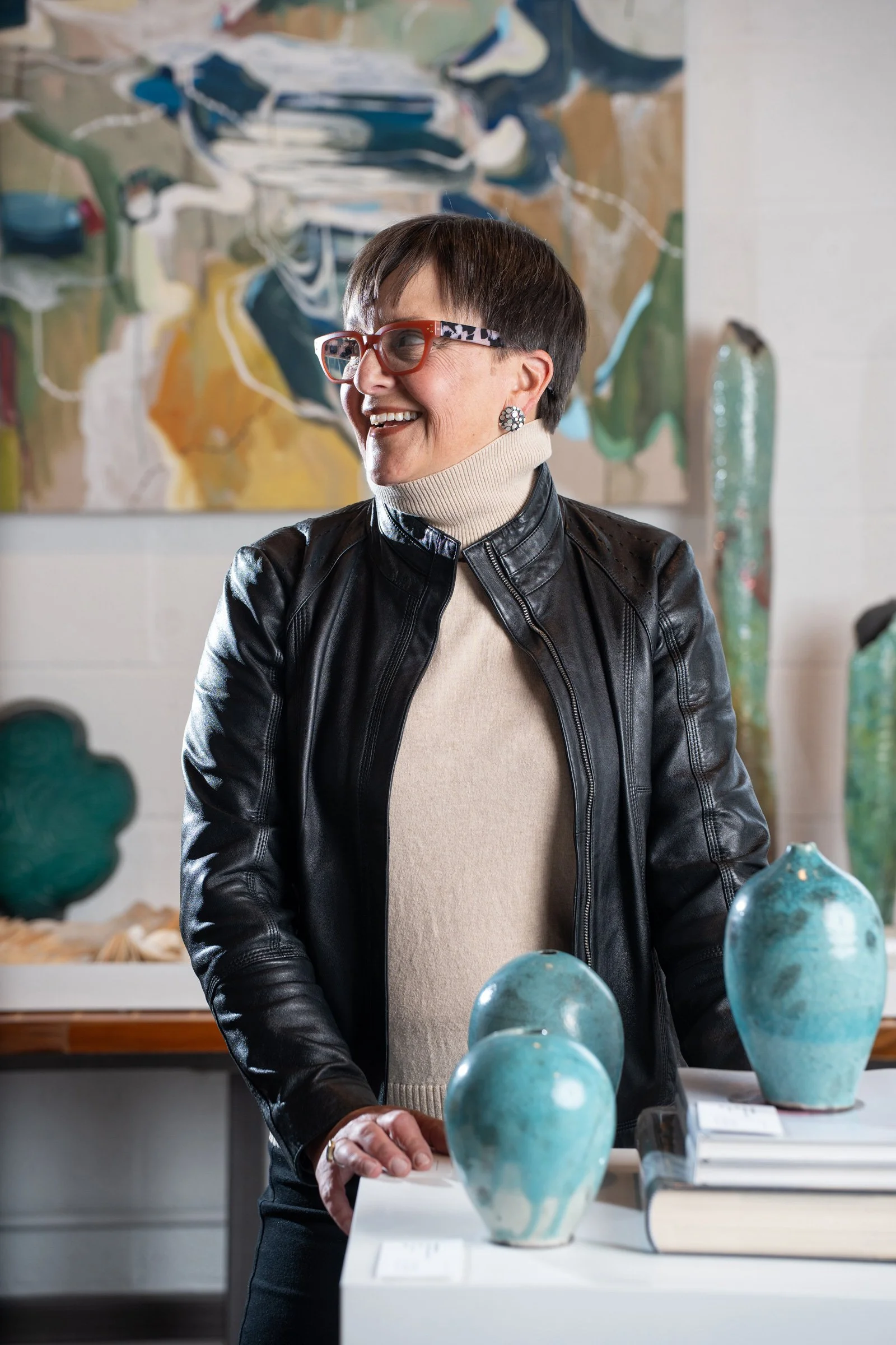 Kelly Taylor, Rocksalt Studio Principal and Creative Director smiling and looking to her left. She is standing in front of a colorful abstract painting, with pottery vases displayed on a table in front of her.