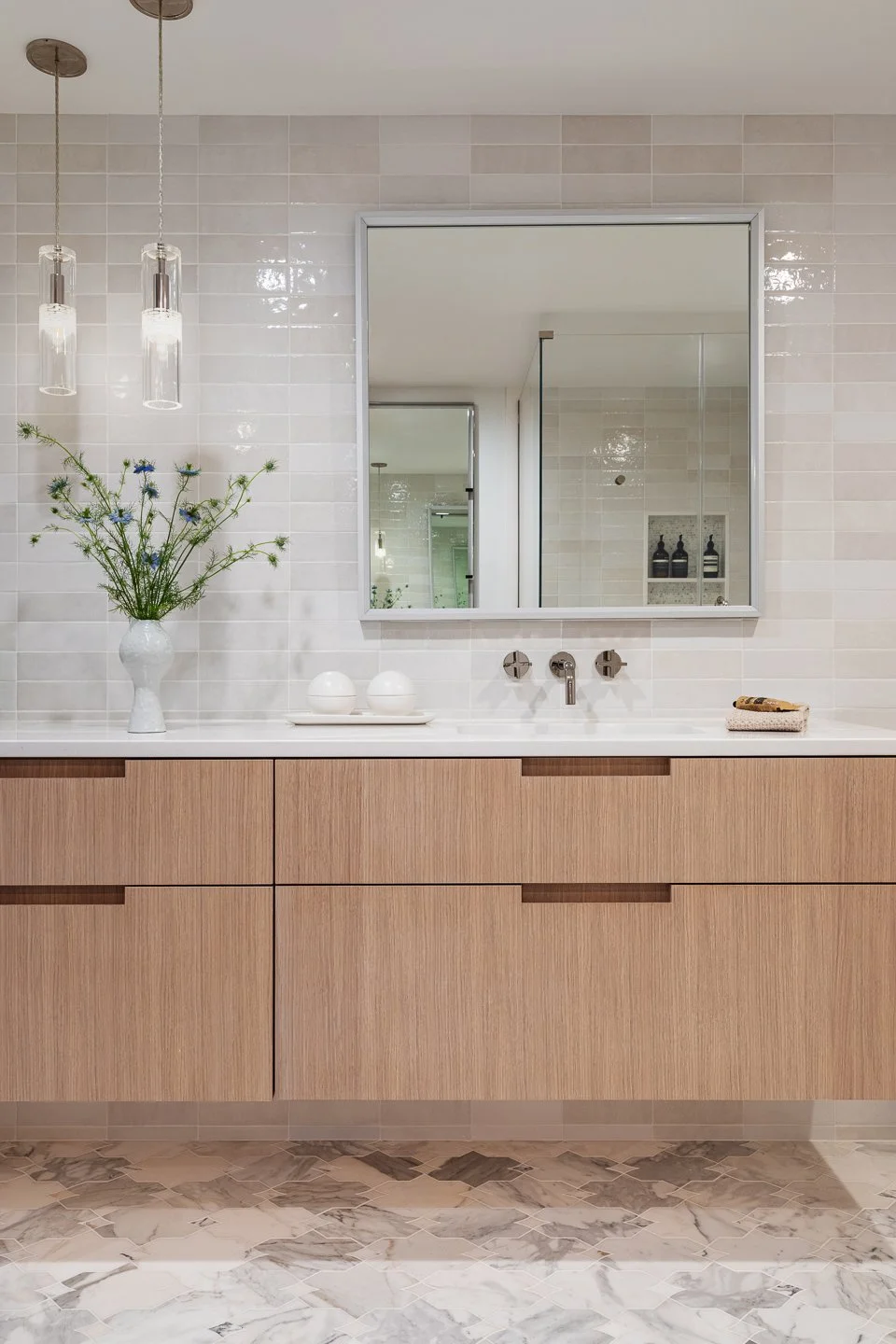 Modern bathroom with a large mirror, wooden vanity, white tiled wall, glass pendant lights, and decorative items like a vase with flowers and small bottles.
