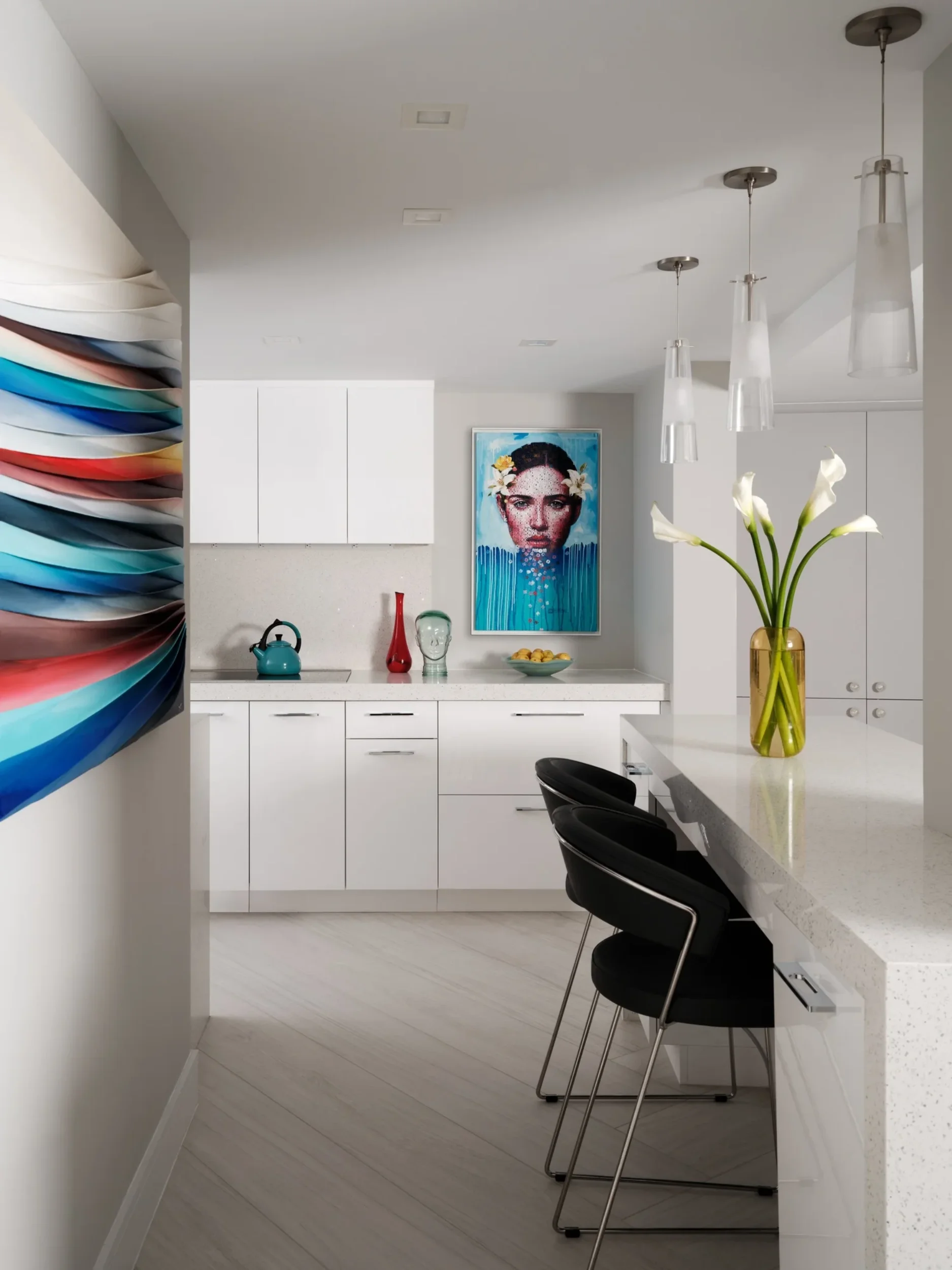 Modern kitchen with white cabinets, a white countertop, black chairs at a bar, a yellow vase with white calla lilies, colorful artwork of a woman with flowers on her head, and hanging pendant lights.