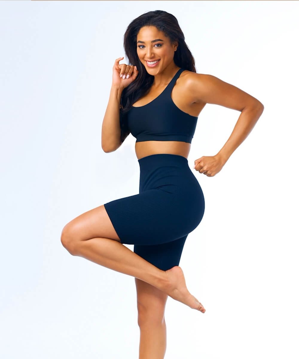 A woman in athletic wear smiles, standing on one leg with the other leg bent and foot on her thigh, and her arms in a pose suggesting strength.