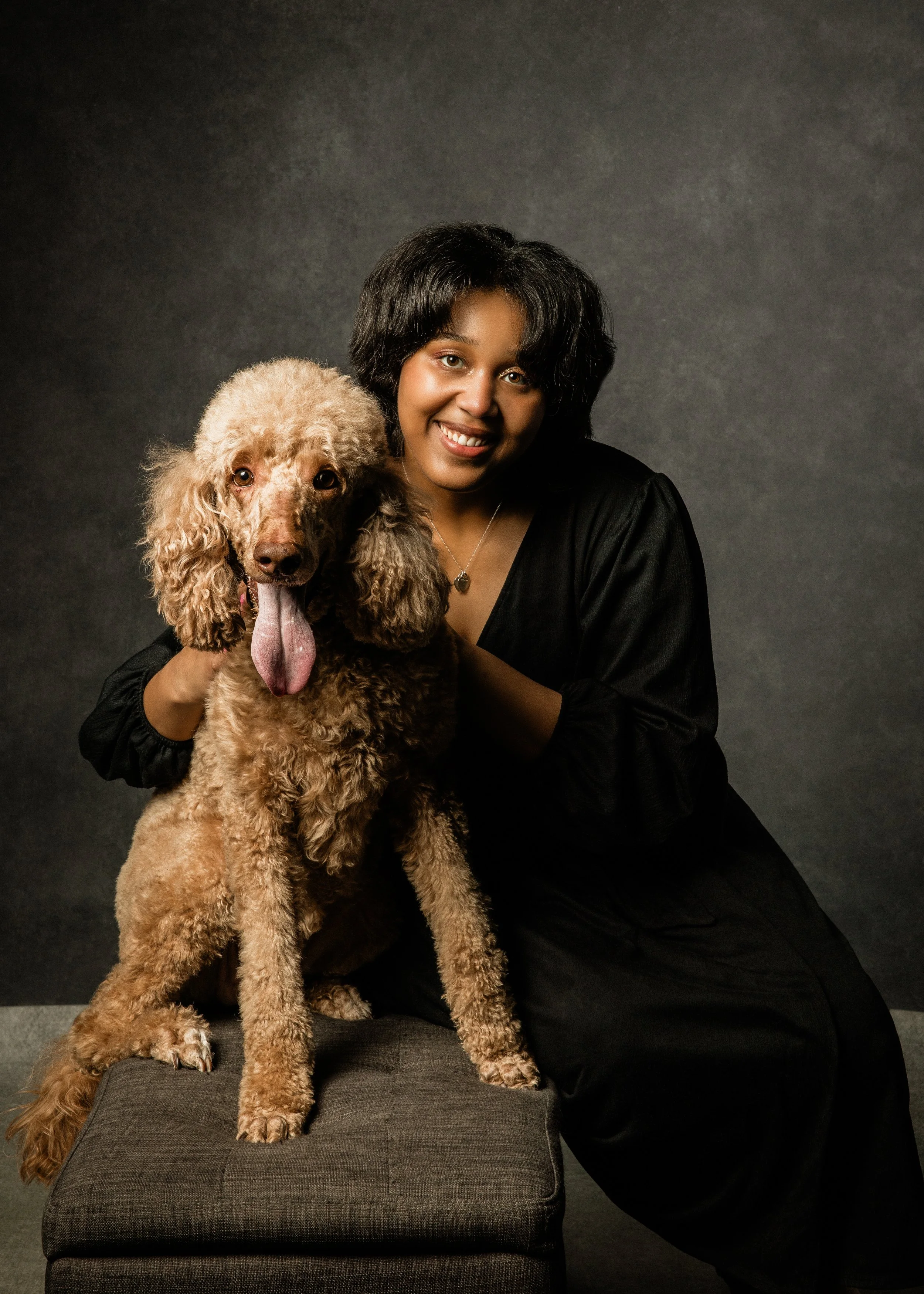 A woman with short black hair and a black top is smiling while holding a large, light brown poodle with curly fur. The woman is sitting next to a gray cushioned ottoman against a dark background.