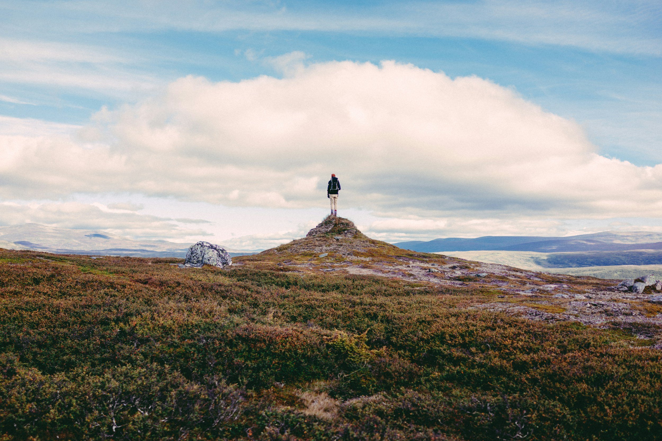 A person standing atop a small hill in a vast, open landscape with a cloudy sky in the background.