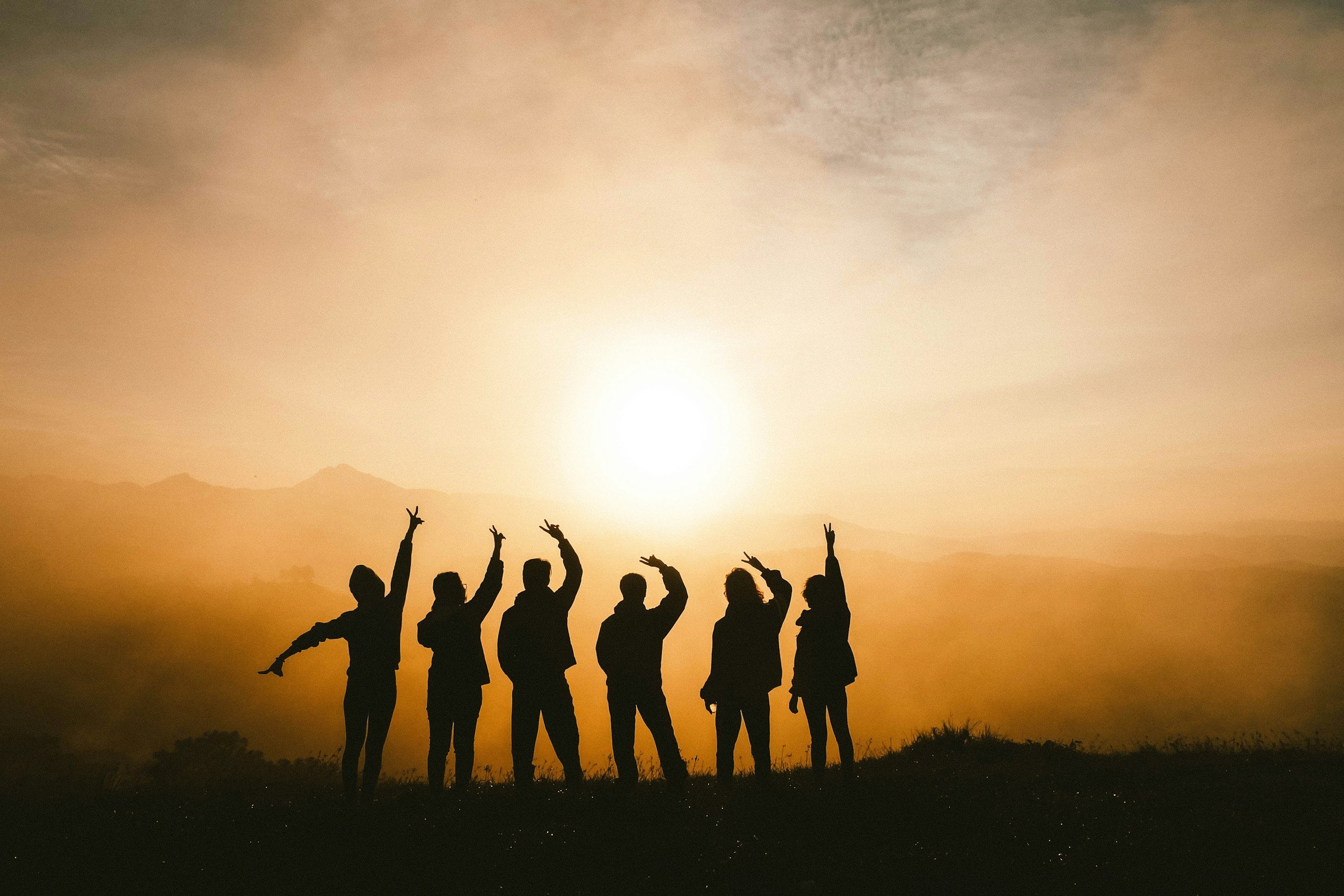 Silhouettes of six people standing outdoors at sunset, some raising their arms, with mountains and a bright sky in the background.