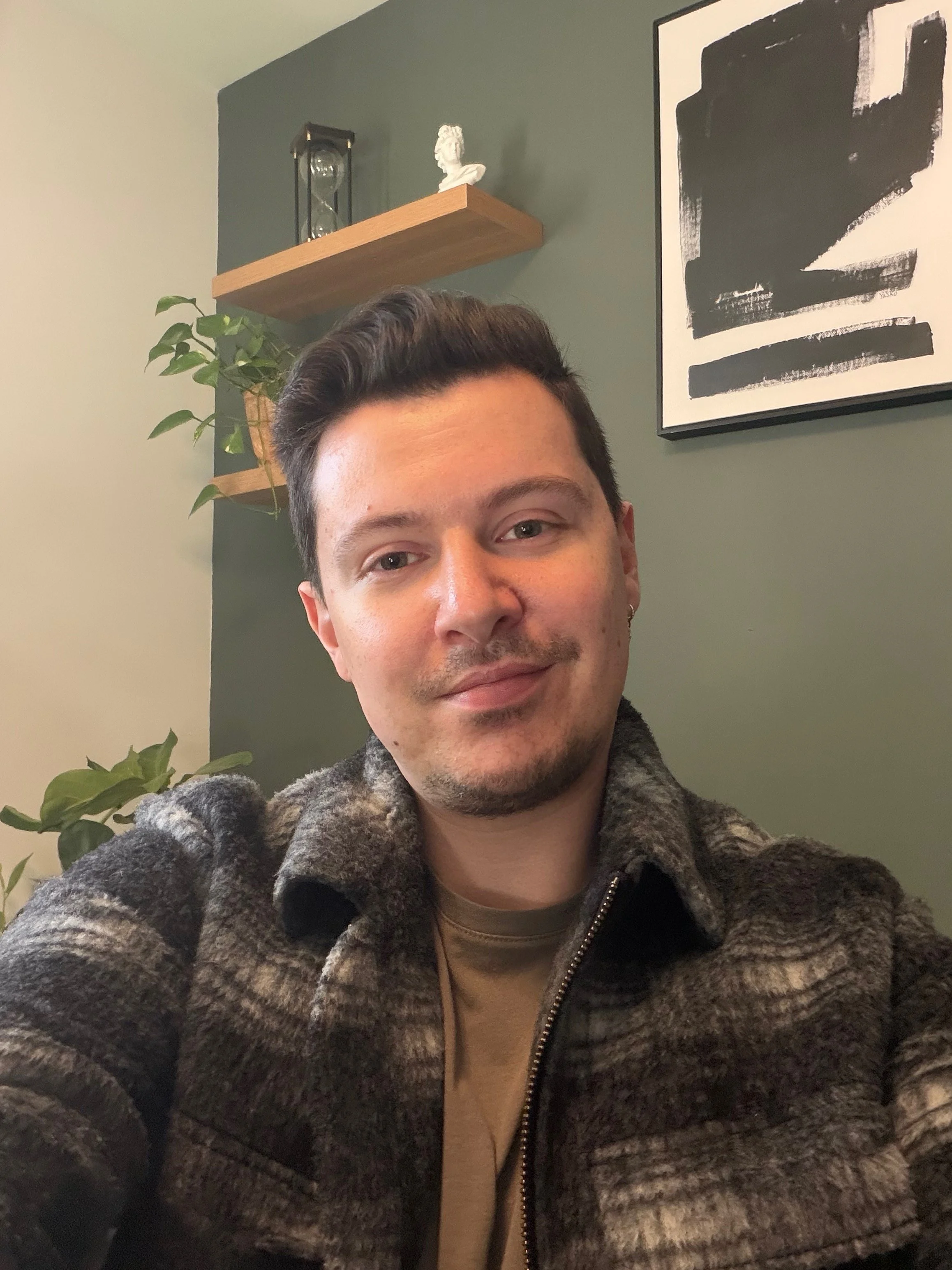 A young man taking a selfie indoors, smiling slightly, with a green wall, decorative plants, and abstract black and white artwork in the background.
