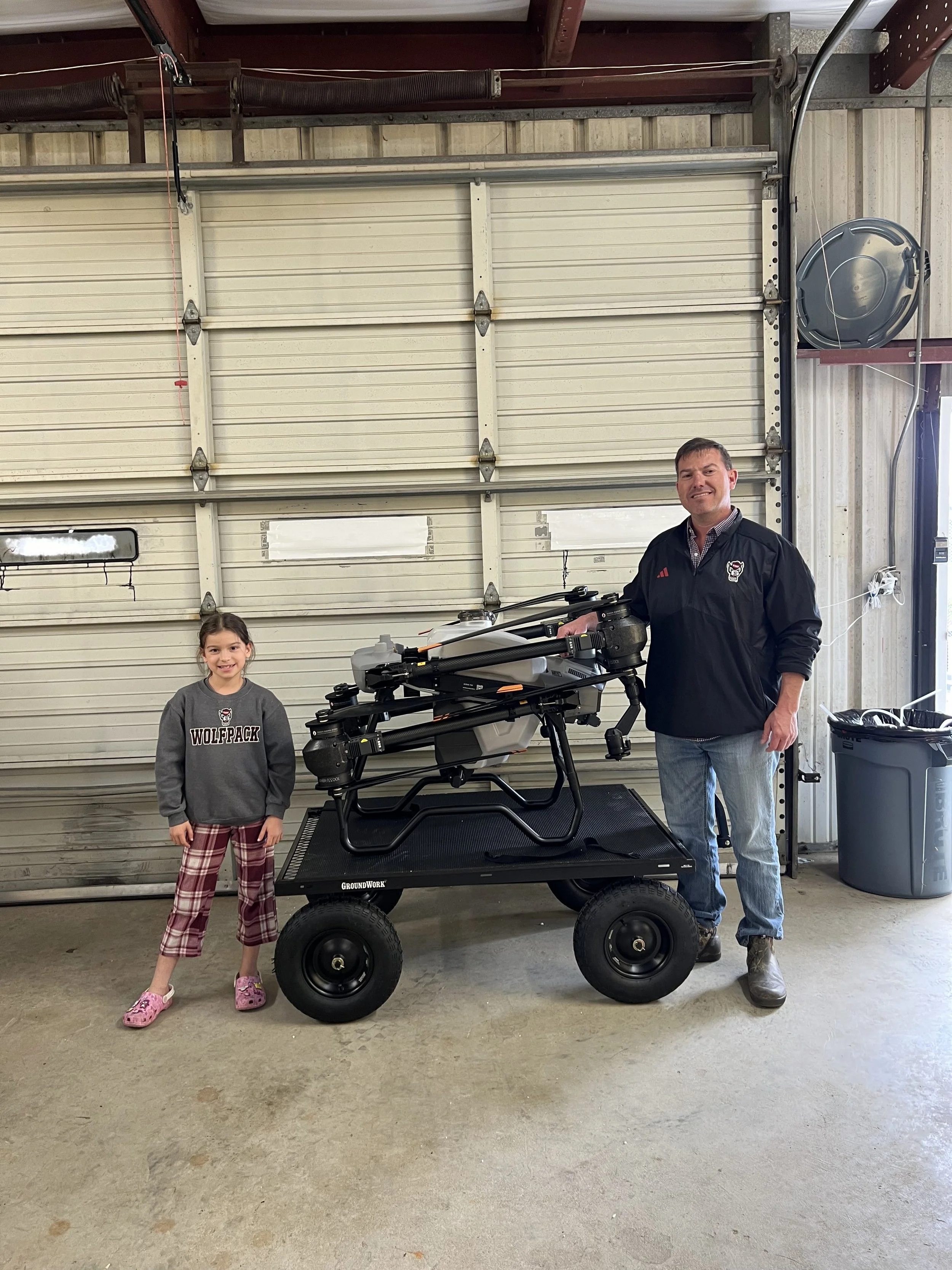 A man and a young girl standing next to a drone on a cart inside a garage with a metal door.