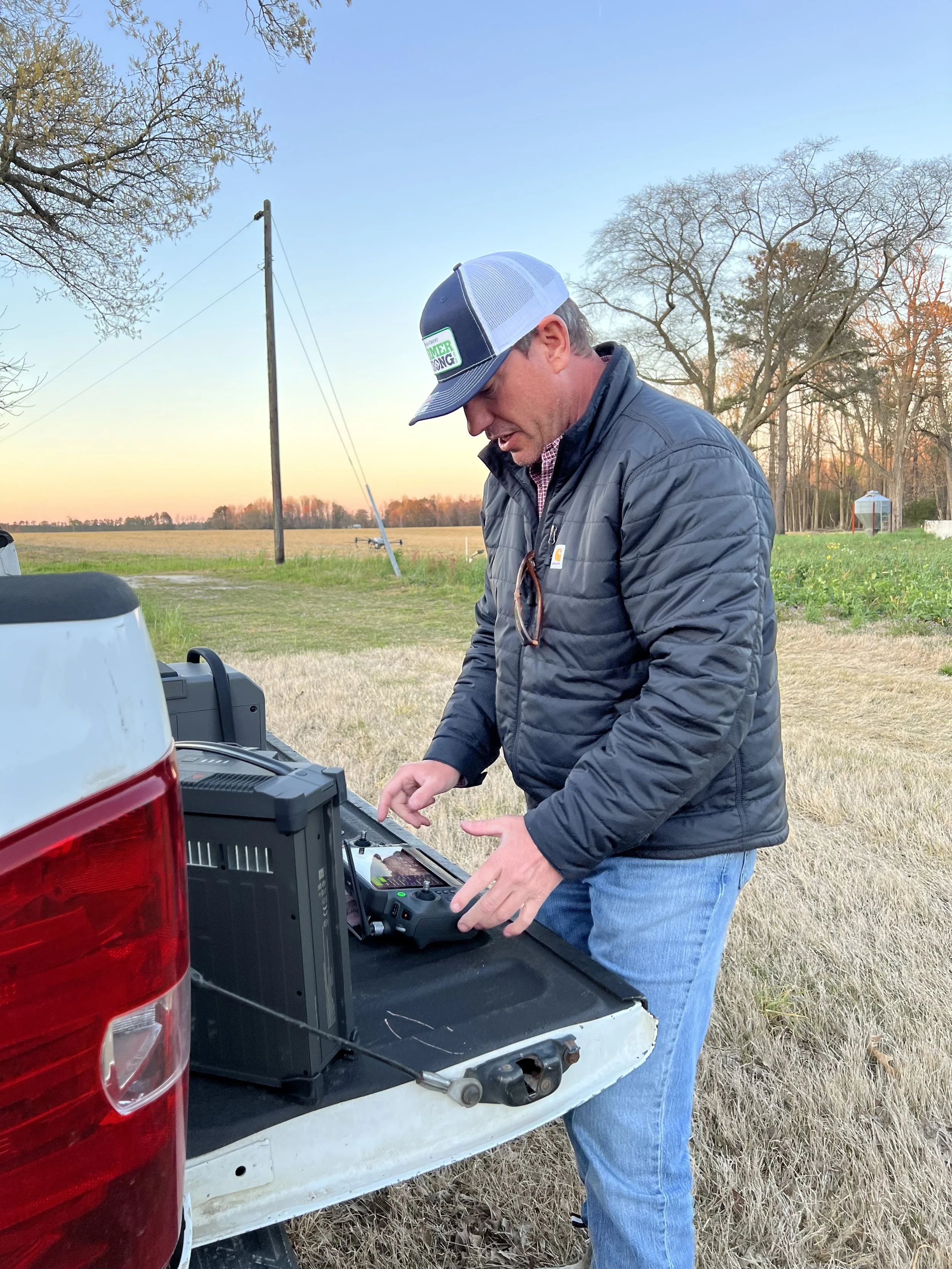 A man wearing a baseball cap, jacket, and jeans operates electronic equipment on the back of a pickup truck in an open grassy field during sunset.
