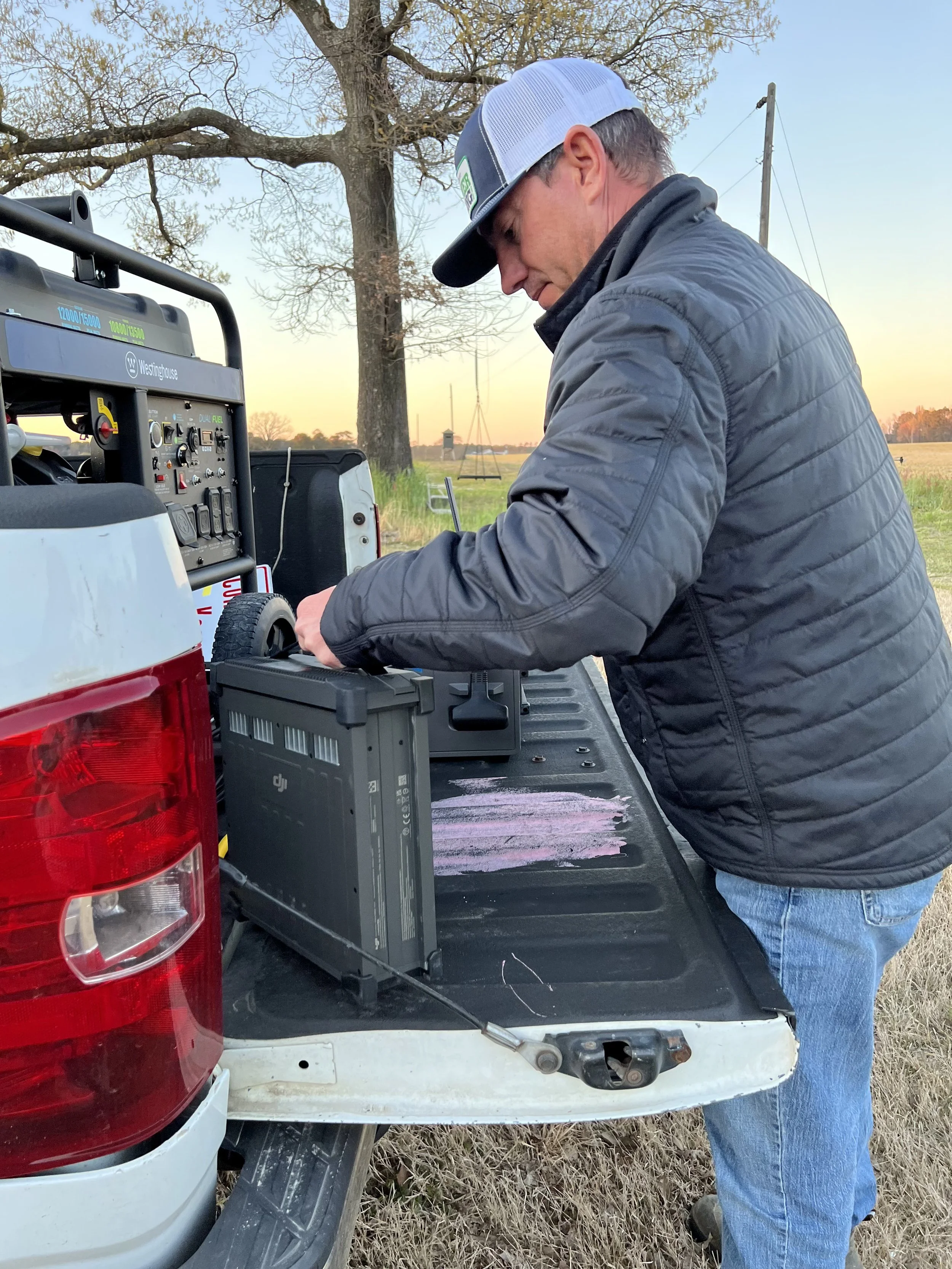 A man wearing a gray jacket, blue jeans, and a trucker hat standing at the back of a pickup truck in an open field, working on a drone battery on the truck bed at sunset with a large tree and power lines in the background.