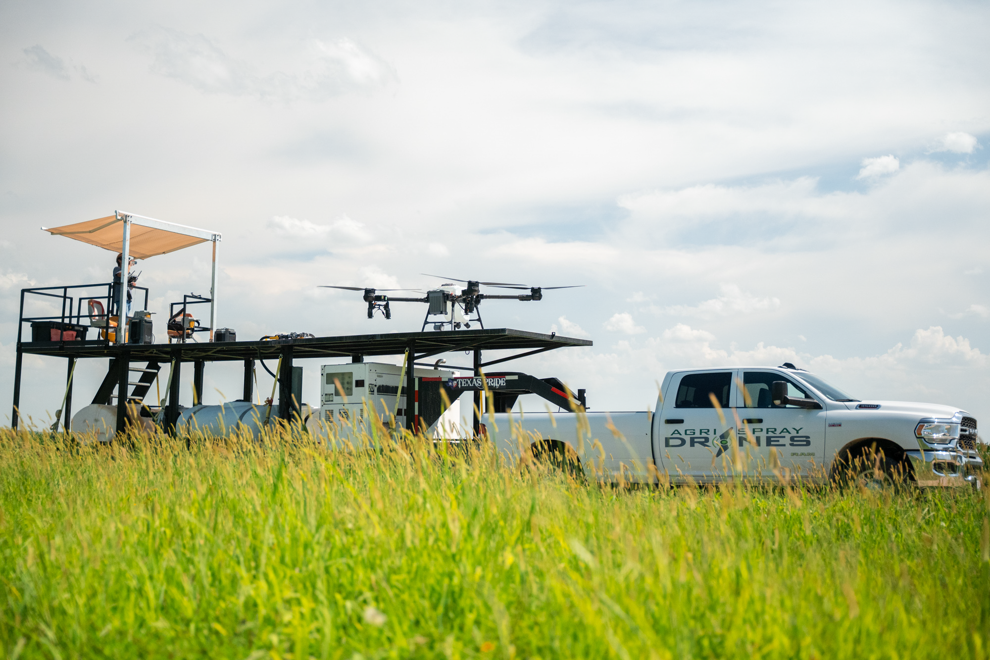 A white pickup truck with 'AGRI SPRAY DROVES' written on the side, parked in a grassy field. On the truck bed, there is a mounted platform with drones equipped with spray tanks and a person operating equipment under a small canopy. Two drones are flying above, and the sky is partly cloudy.