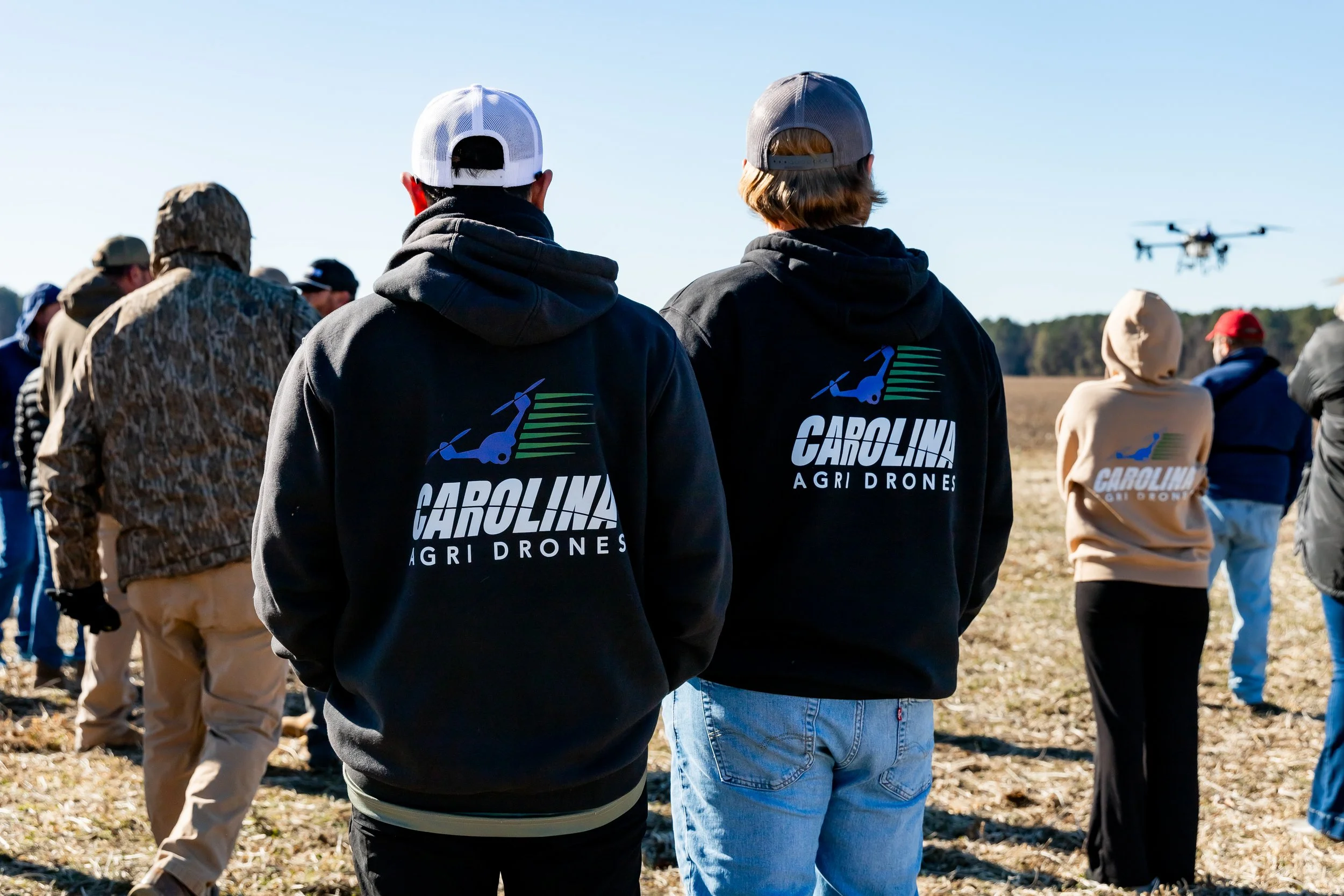 Group of people standing outdoors in a field, some wearing Carolina Agri Drones hoodies, observing a drone flying in the sky on a clear day.