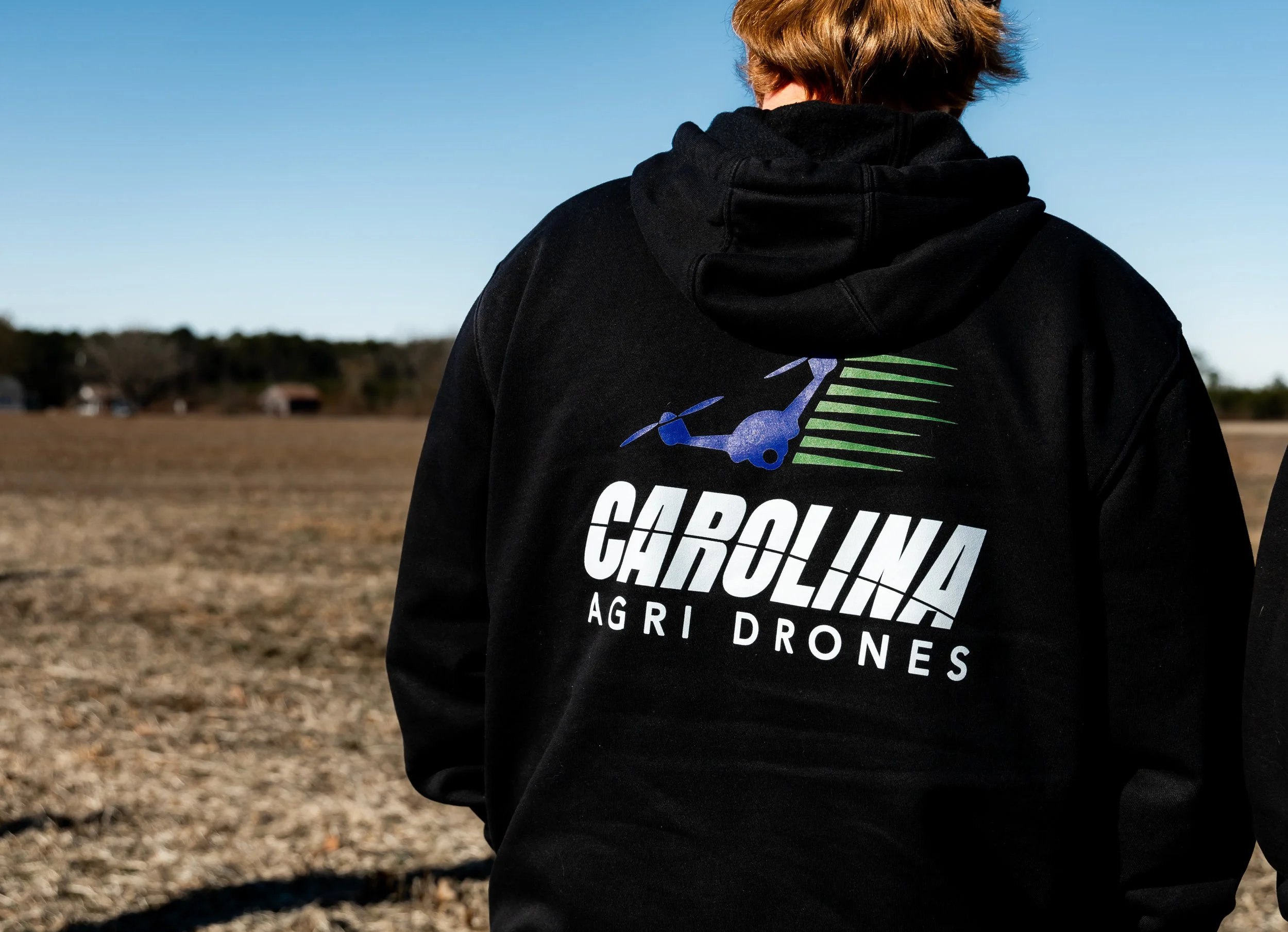 Person standing outdoors in a field wearing a black hoodie with 'Carolina Agri Drones' logo on the back, featuring a drone and text.