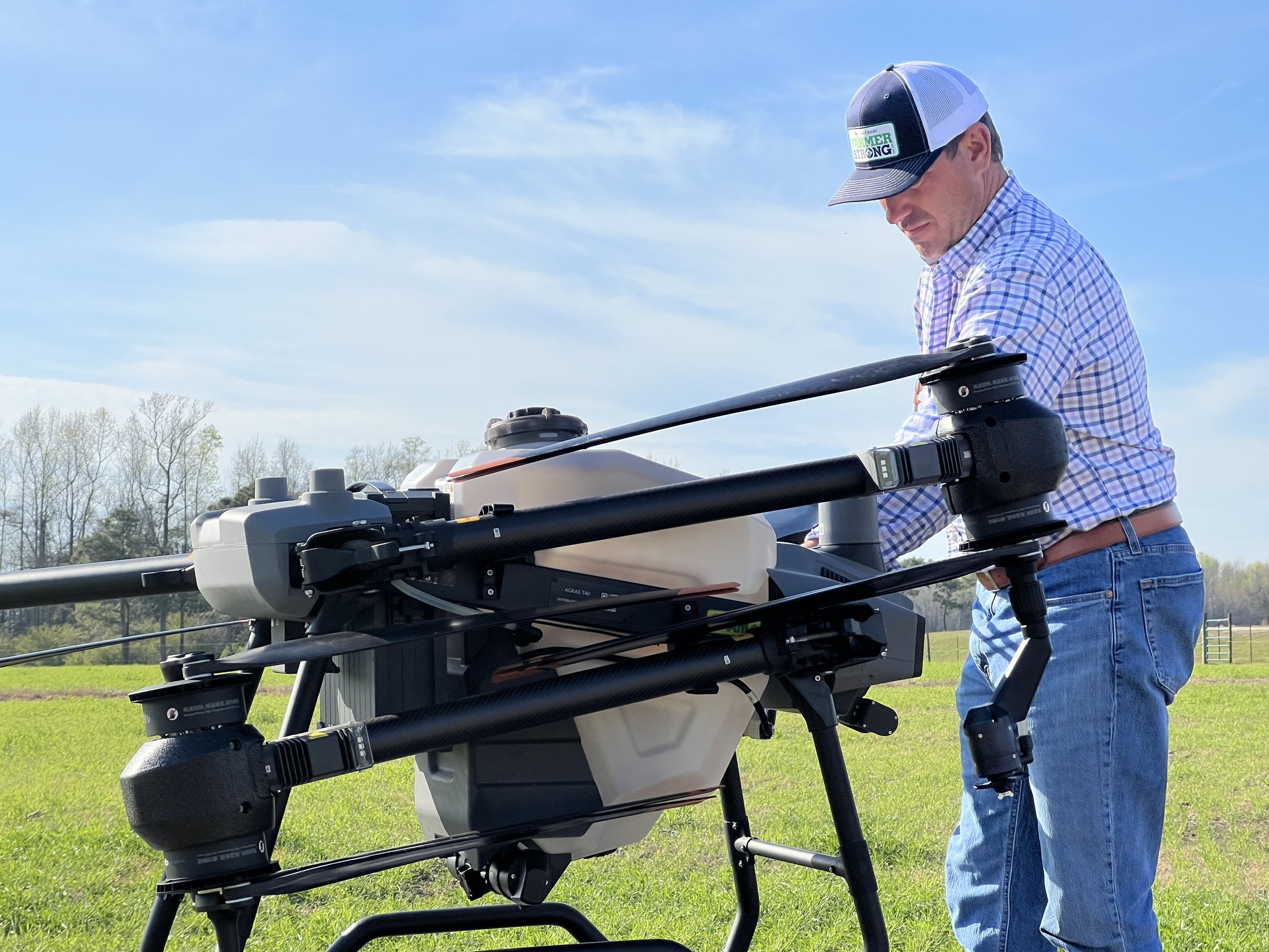 A man in a plaid shirt, blue jeans, and a baseball cap with the words 'GROWER STRONG' is operating a large agricultural drone in a grassy field with trees and a blue sky in the background.