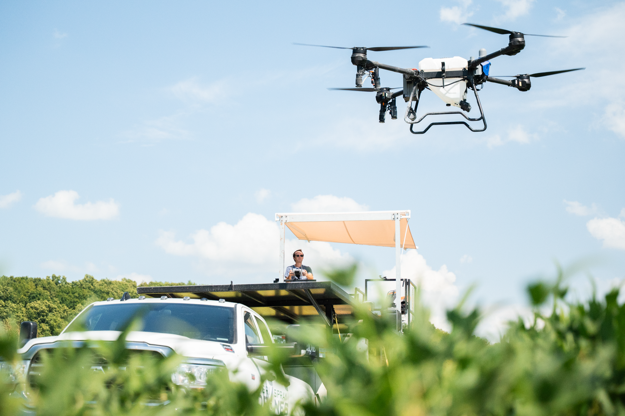 A drone flying above a truck with a man controlling it, in a field with a blue sky and trees in the background.