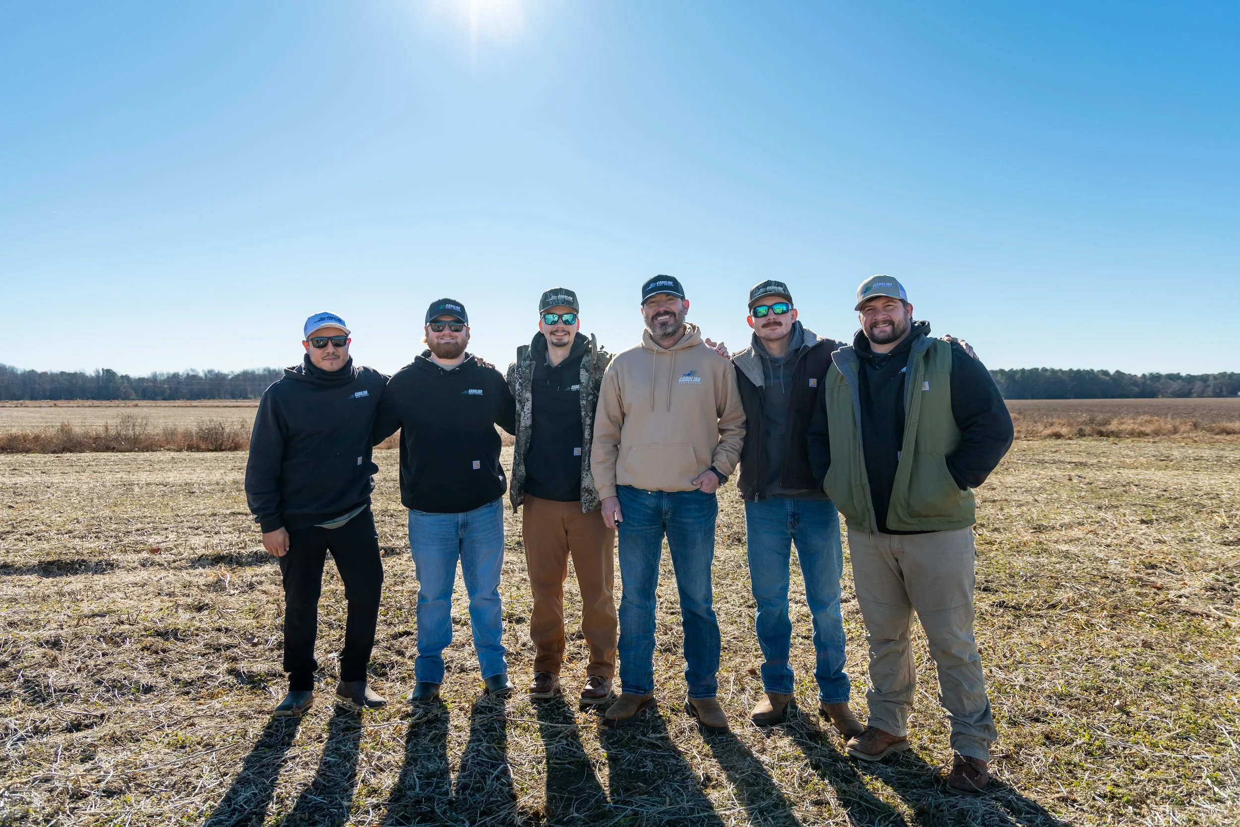 Six men standing together outdoors in a field on a sunny day, dressed in casual outdoor clothing and hats.
