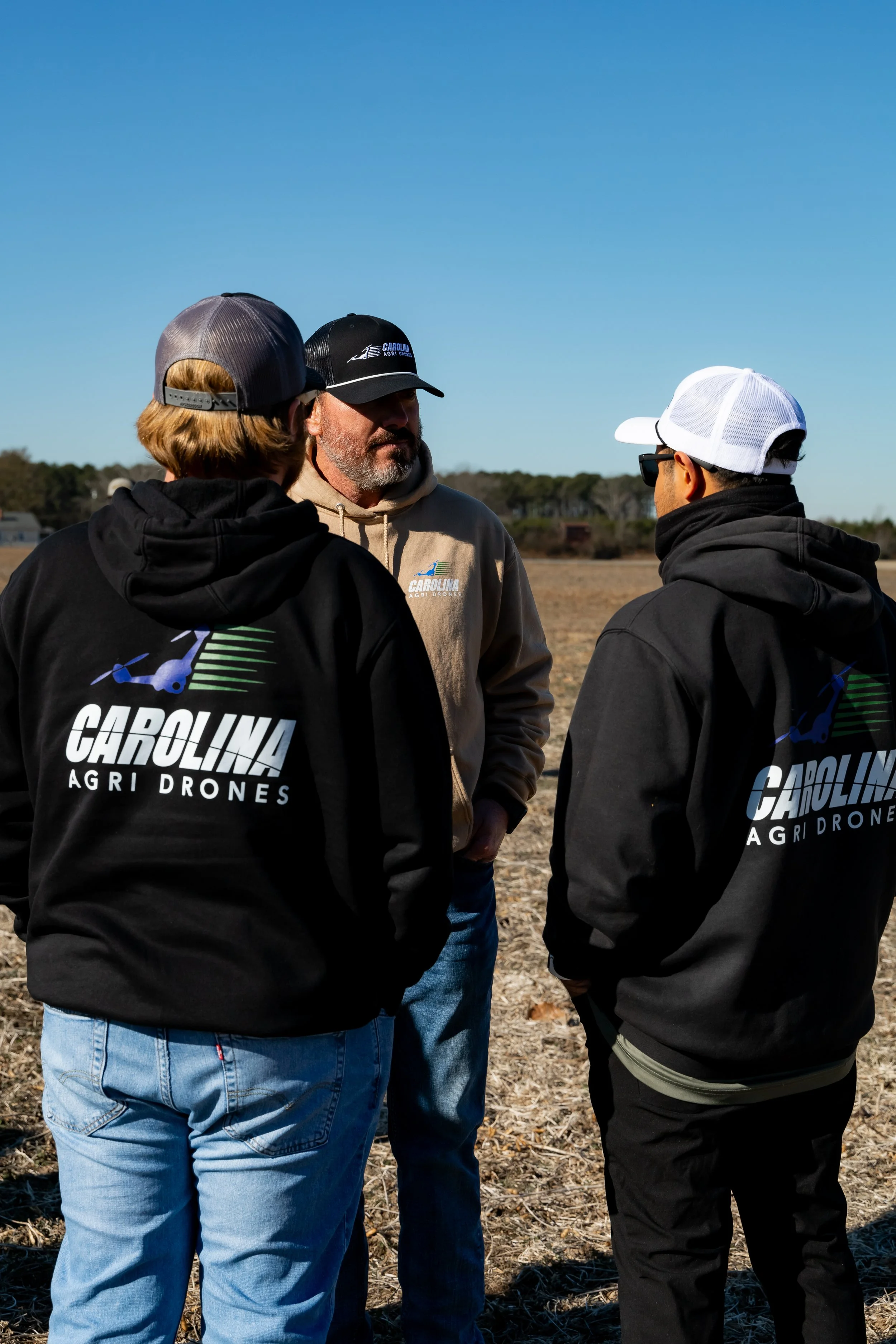 Three men standing outdoors in a field under a clear blue sky, engaged in conversation. Two are wearing black hoodies with the logo 'Carolina Agri Drones'.