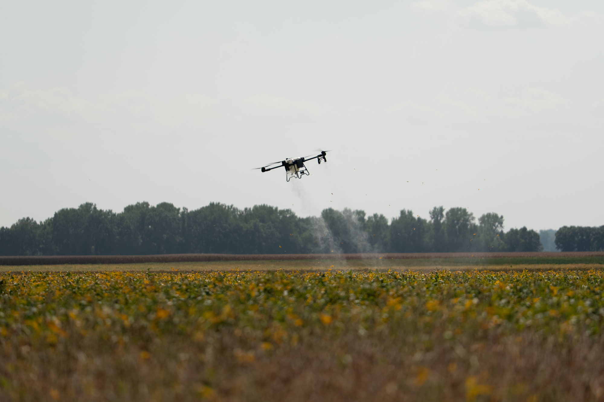 A drone flying over a farm field, dispersing seeds or chemicals, with trees and a partly cloudy sky in the background.